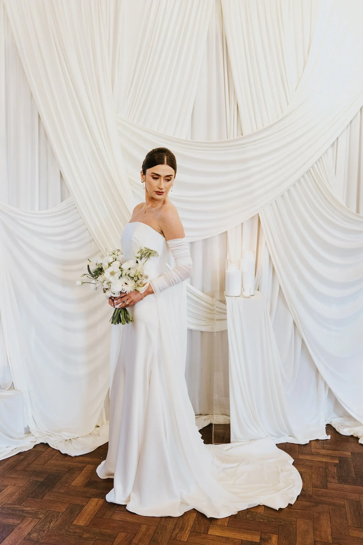 A bride in a white wedding dress holding a bouquet of white flowers standing in front of a cream-colored, draped fabric backdrop.