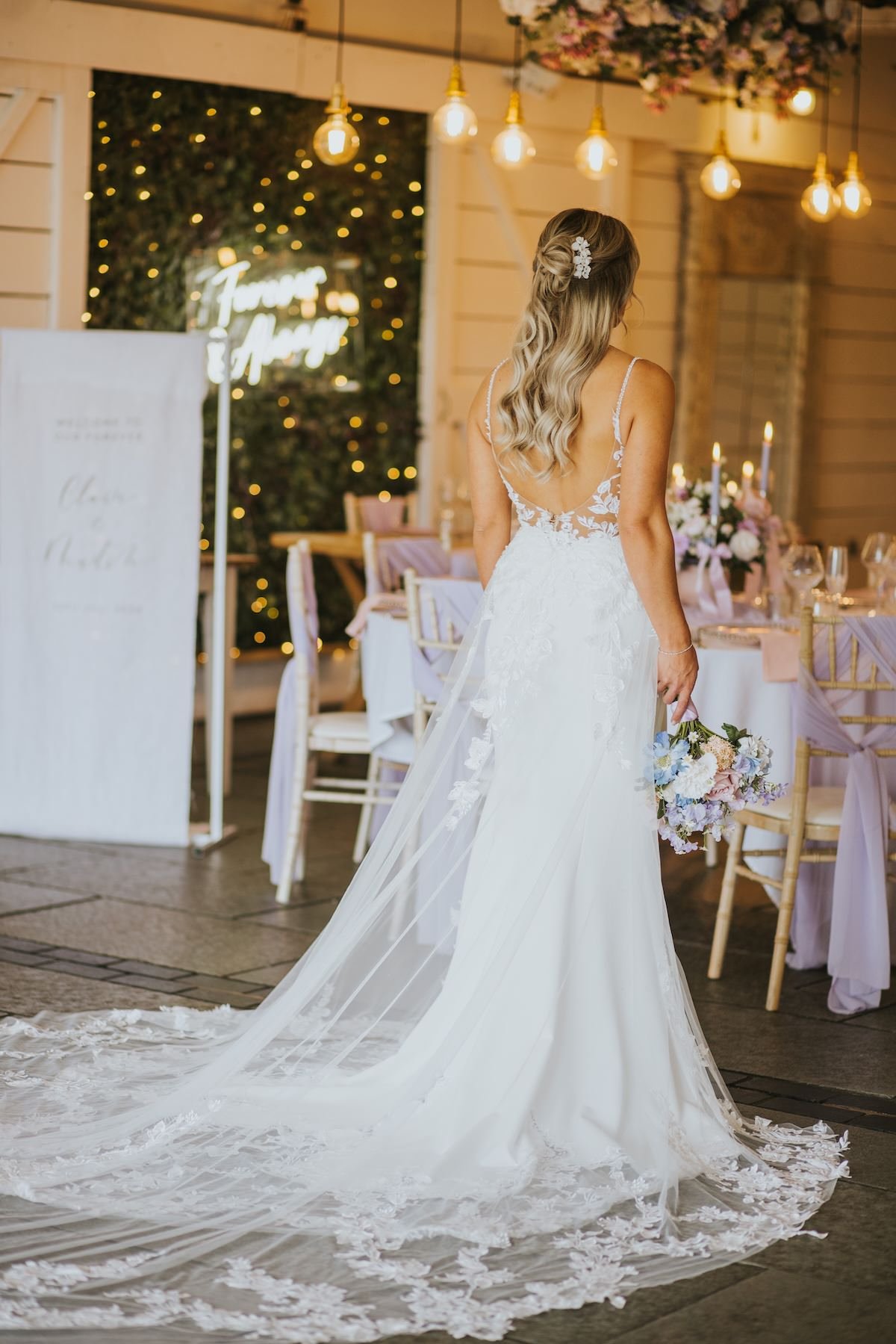 A bride in a white wedding gown with lace details and a long train, holding a bouquet of pastel-colored flowers, standing in a decorated reception area with tables, chairs, floral arrangements, candles, and warm hanging lights.