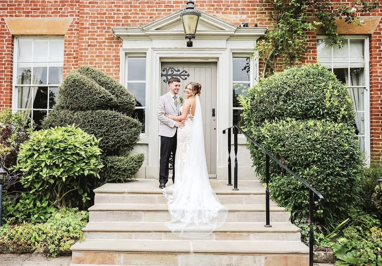A newlywed couple standing on steps in front of a brick house with large windows and bushes, smiling at each other.