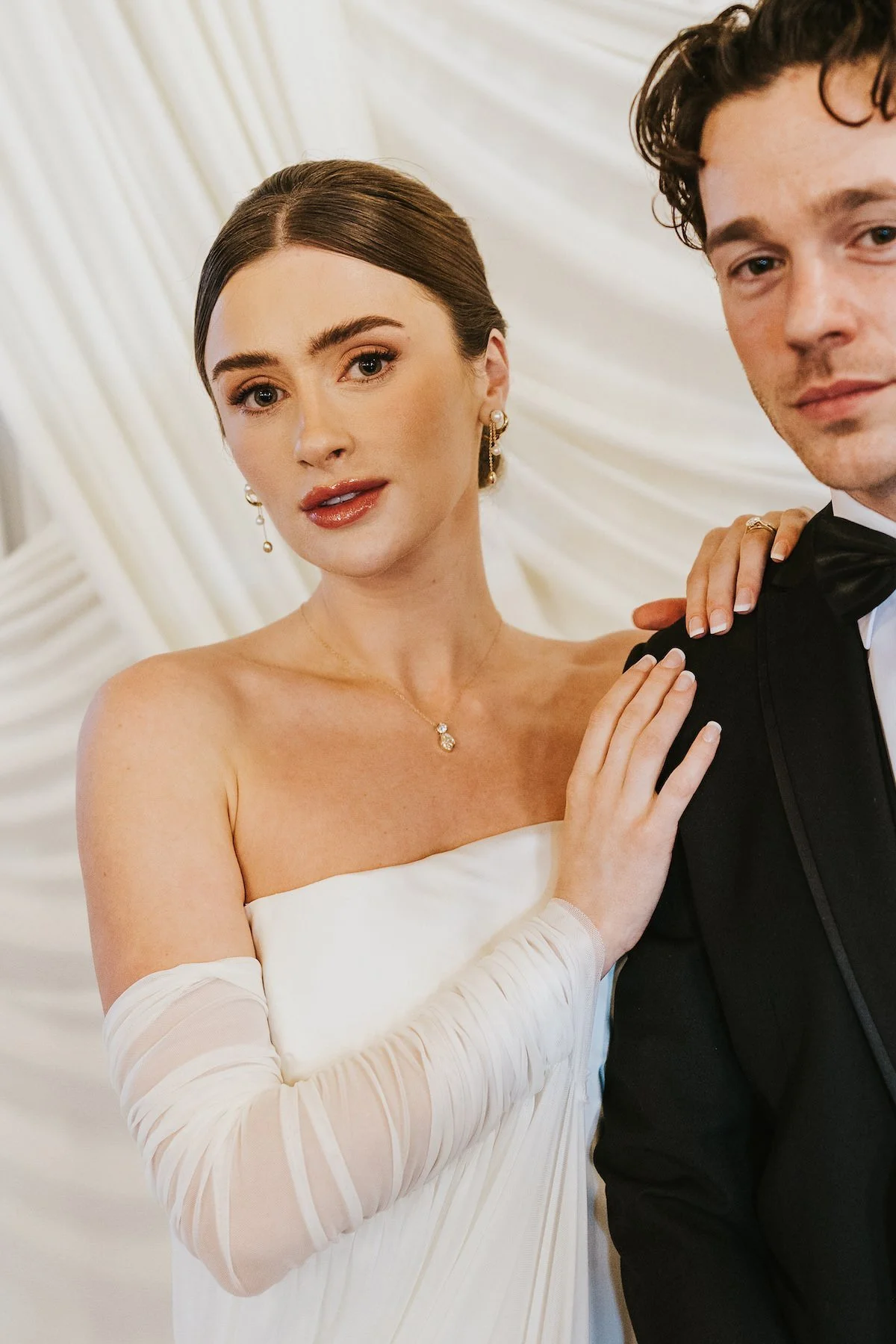 A bride and groom in formal wedding attire close up portrait, with the bride wearing a white off-shoulder dress and jewelry, and the groom in a black tuxedo with a bow tie, posing together at their wedding reception.