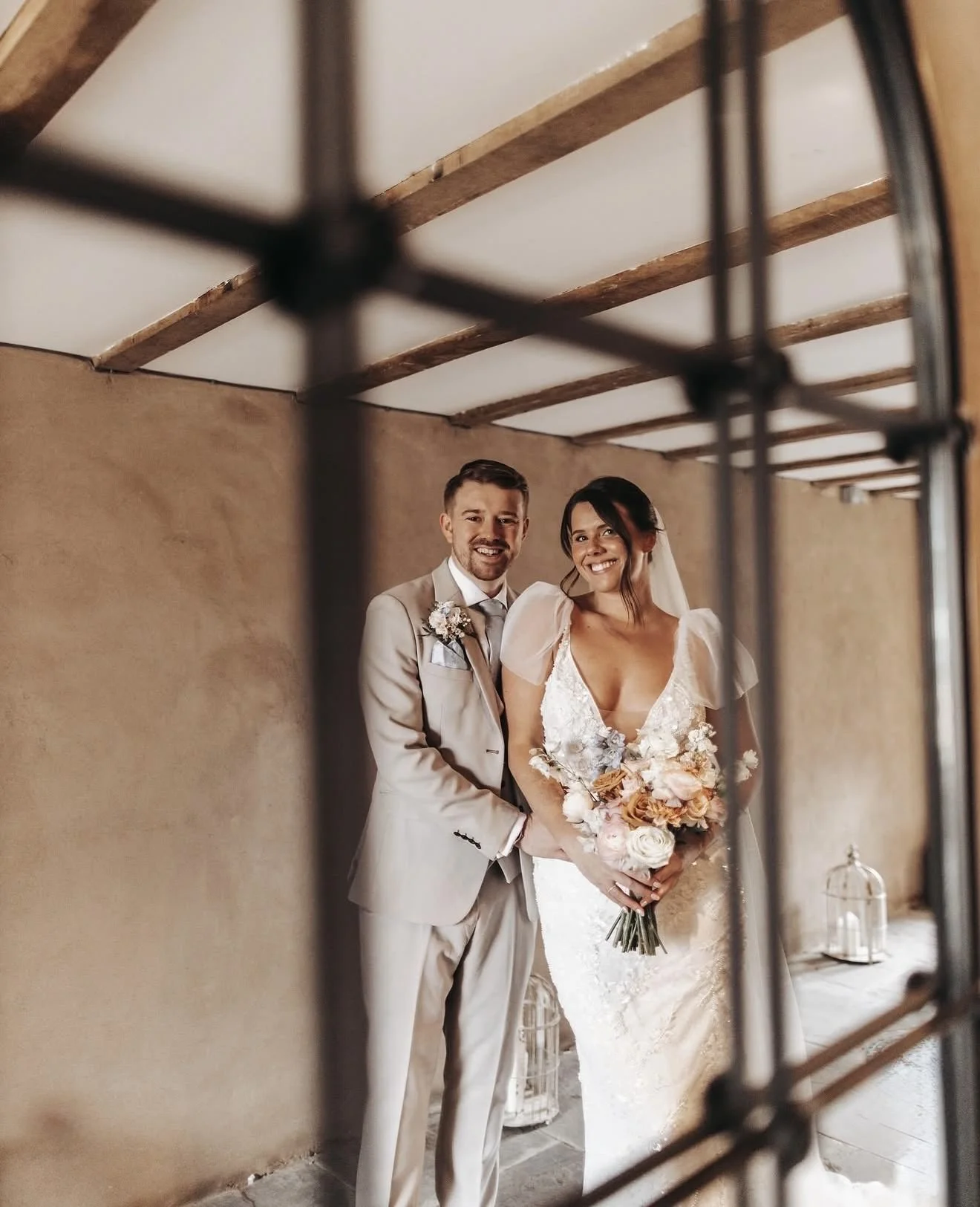 A happy bride and groom are smiling for a photo during their wedding, holding a bouquet of flowers, through a metal grid or cage.