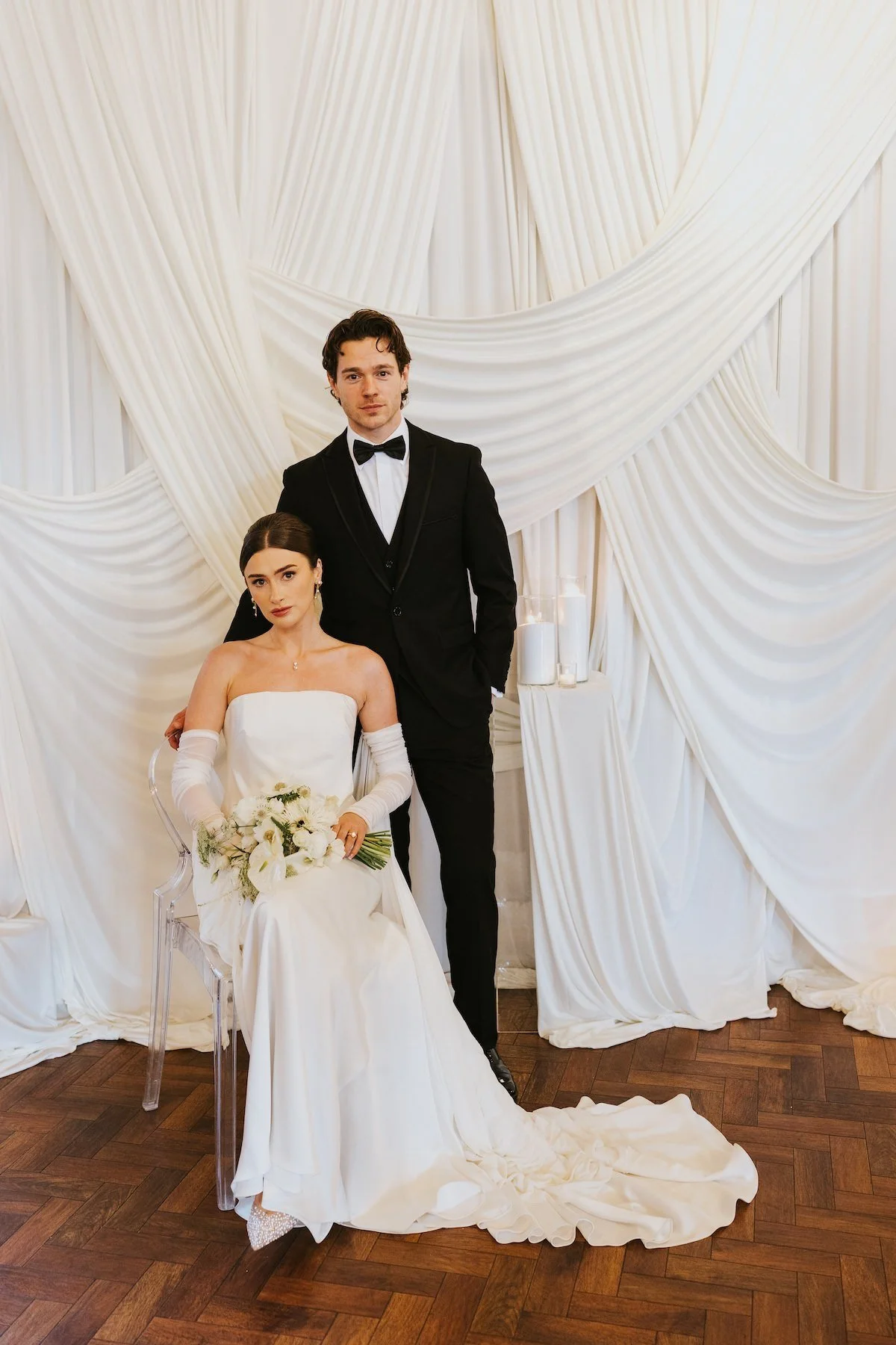 A bride and groom posing together at their wedding. The bride is seated in a transparent chair, wearing a white strapless wedding gown with long gloves and holding a bouquet of white flowers. The groom is standing beside her, dressed in a black tuxed