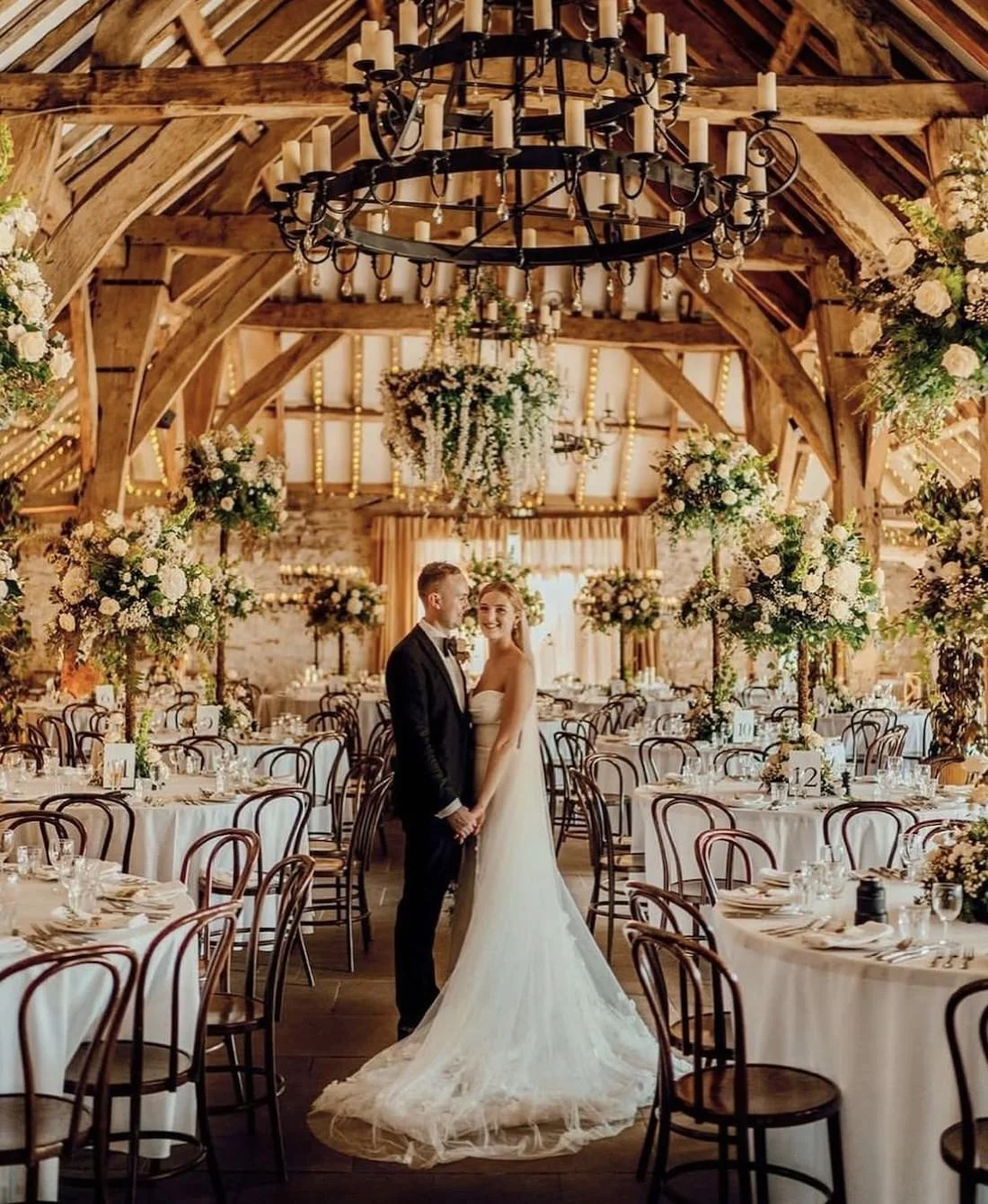 A bride and groom standing and holding hands in a beautifully decorated wedding reception hall with floral arrangements, wooden beams, and a chandelier overhead.