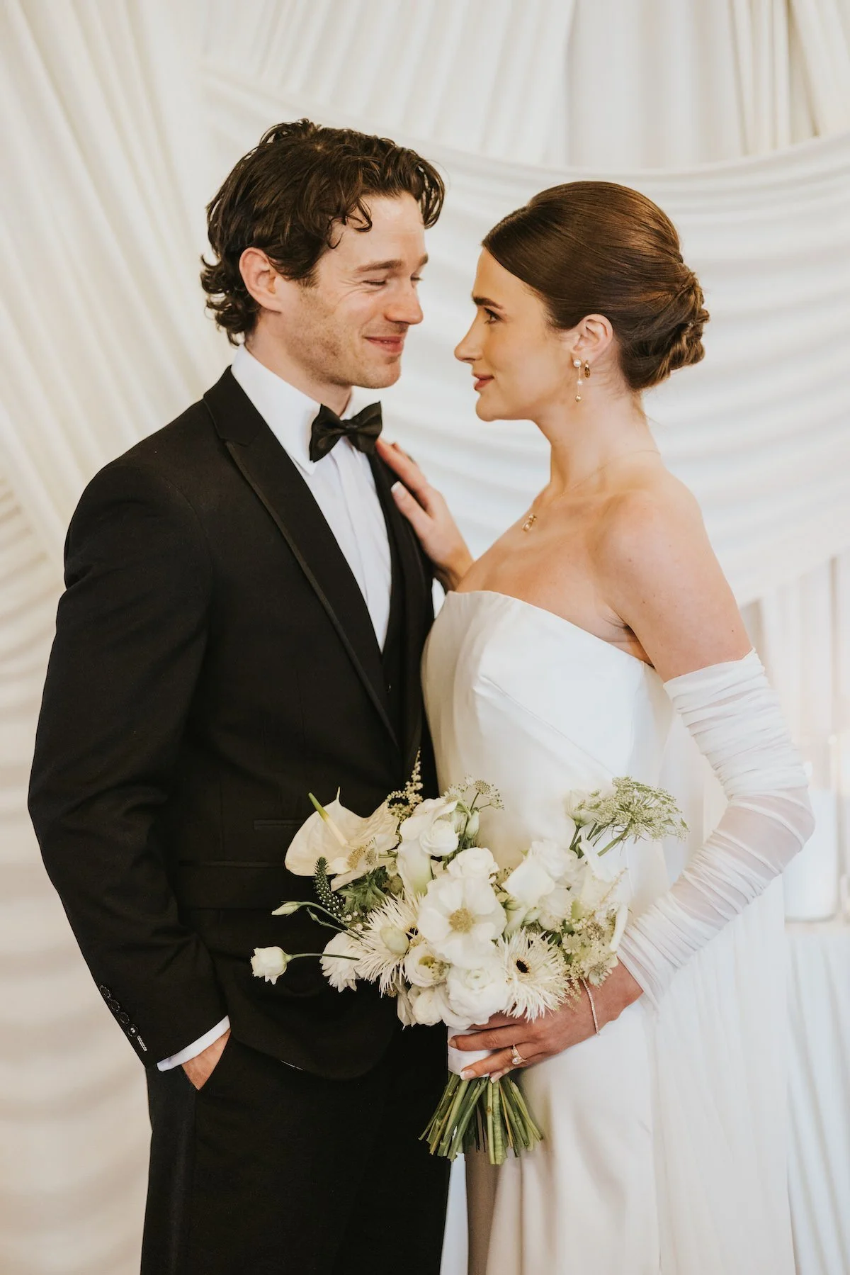A bride and groom on their wedding day, standing close and gazing at each other, with the bride holding a bouquet of white flowers.