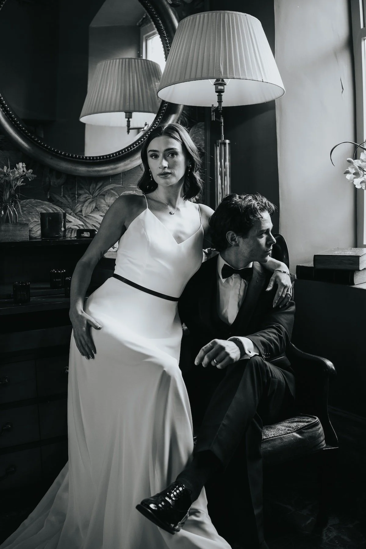 A black and white photo of a woman in a white dress standing next to a seated man in a tuxedo, in a dimly lit room with a large mirror, table, lamps, and flowers in the background.