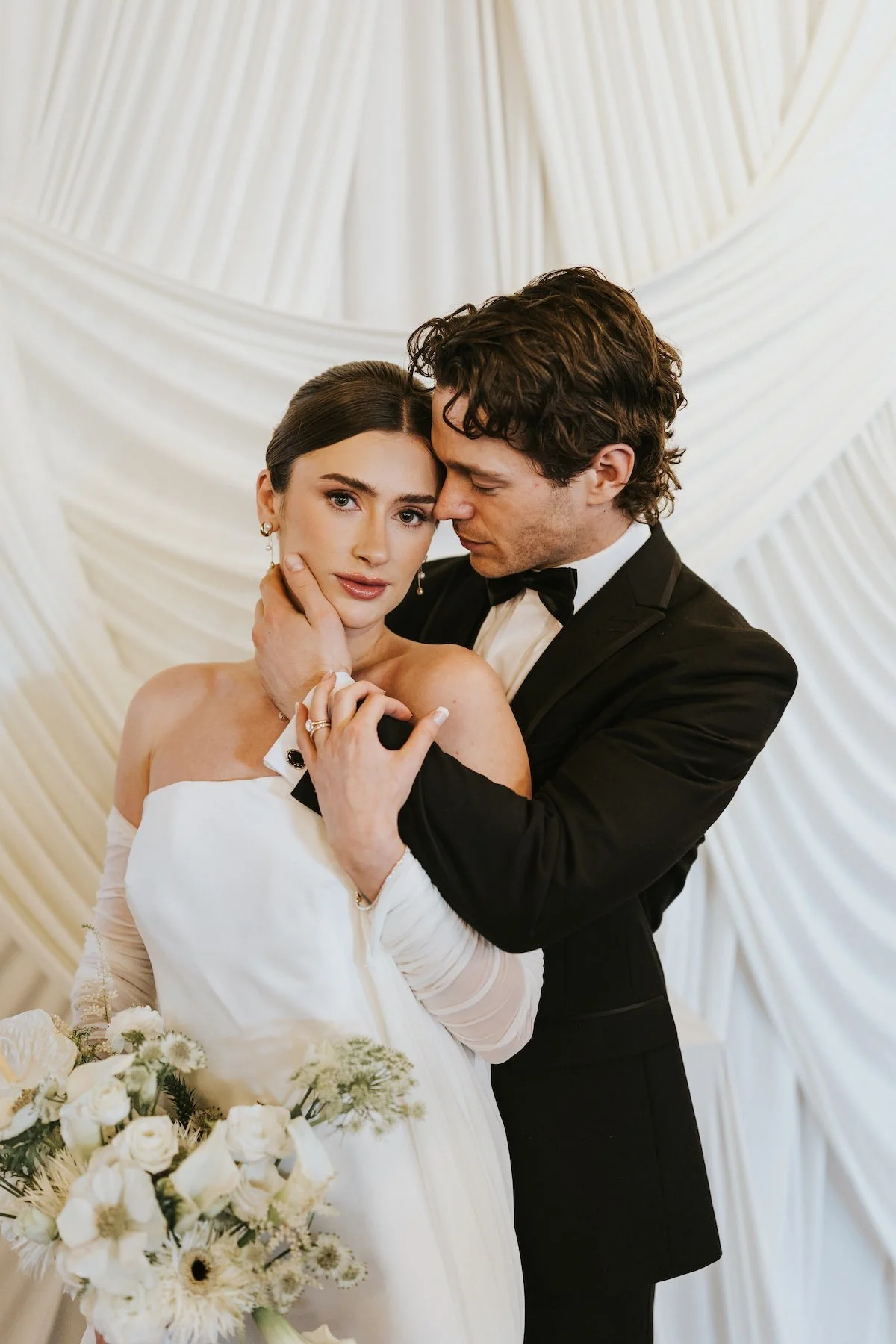 A bride and groom in wedding attire embracing in front of a draped cream-colored backdrop. The bride is holding a bouquet of white flowers and roses, and they are looking into each other's eyes.