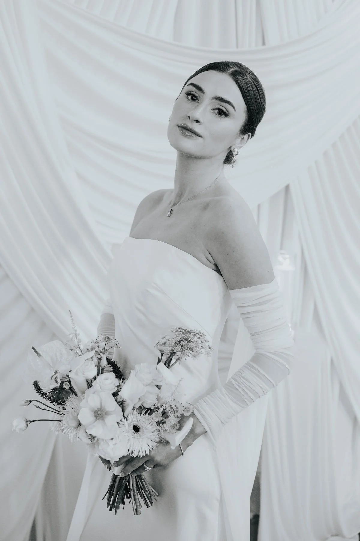 A woman in a wedding dress holding a bouquet of flowers, standing in front of draped fabric backdrop.