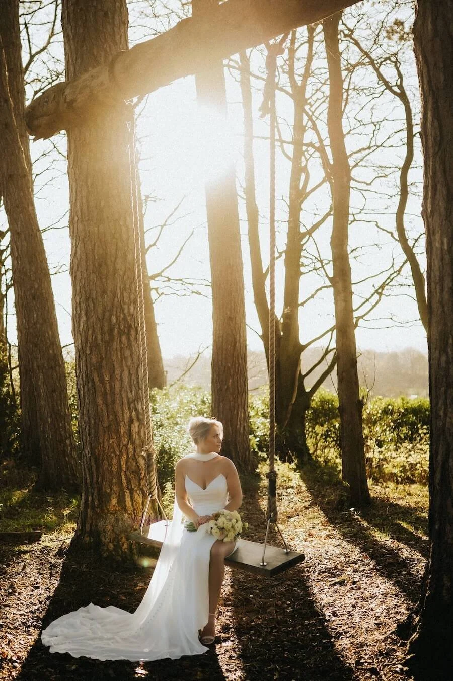 A bride in a white wedding dress and holding a bouquet of white roses sitting on a wooden swing hanging between trees in a forest with sunlight streaming through the branches.