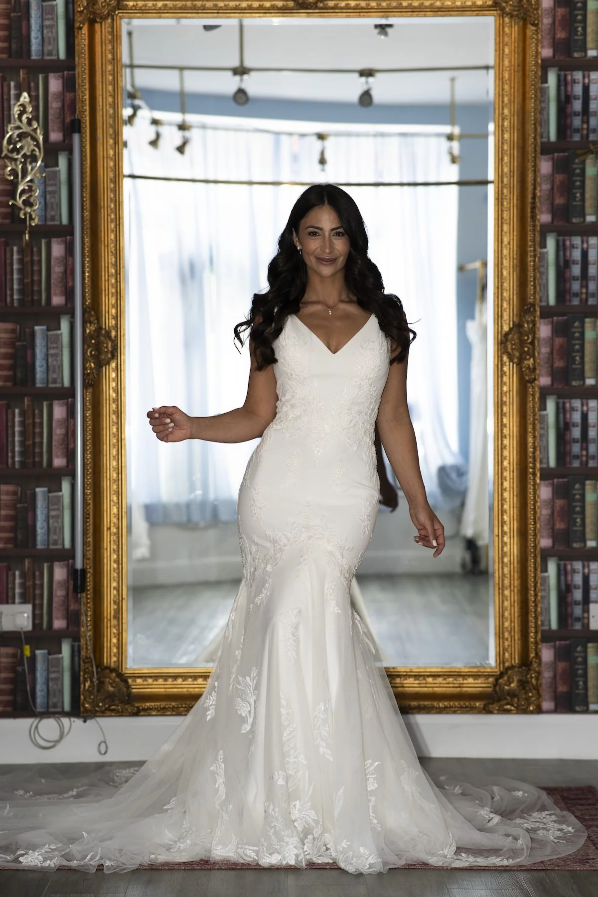 A woman in a white wedding dress standing in front of a large ornate gold mirror. She has long dark curly hair and is smiling, with a background of windows and bookshelves.
