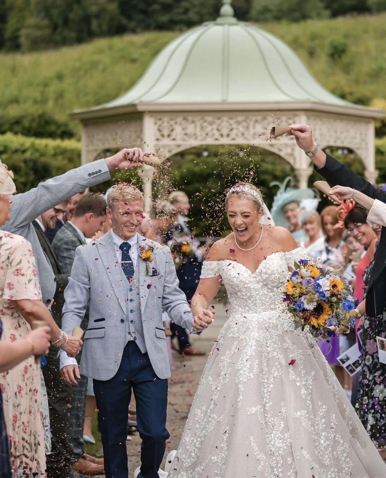 A bride and groom celebrate their wedding with guests throwing confetti in front of a gazebo, surrounded by greenery.