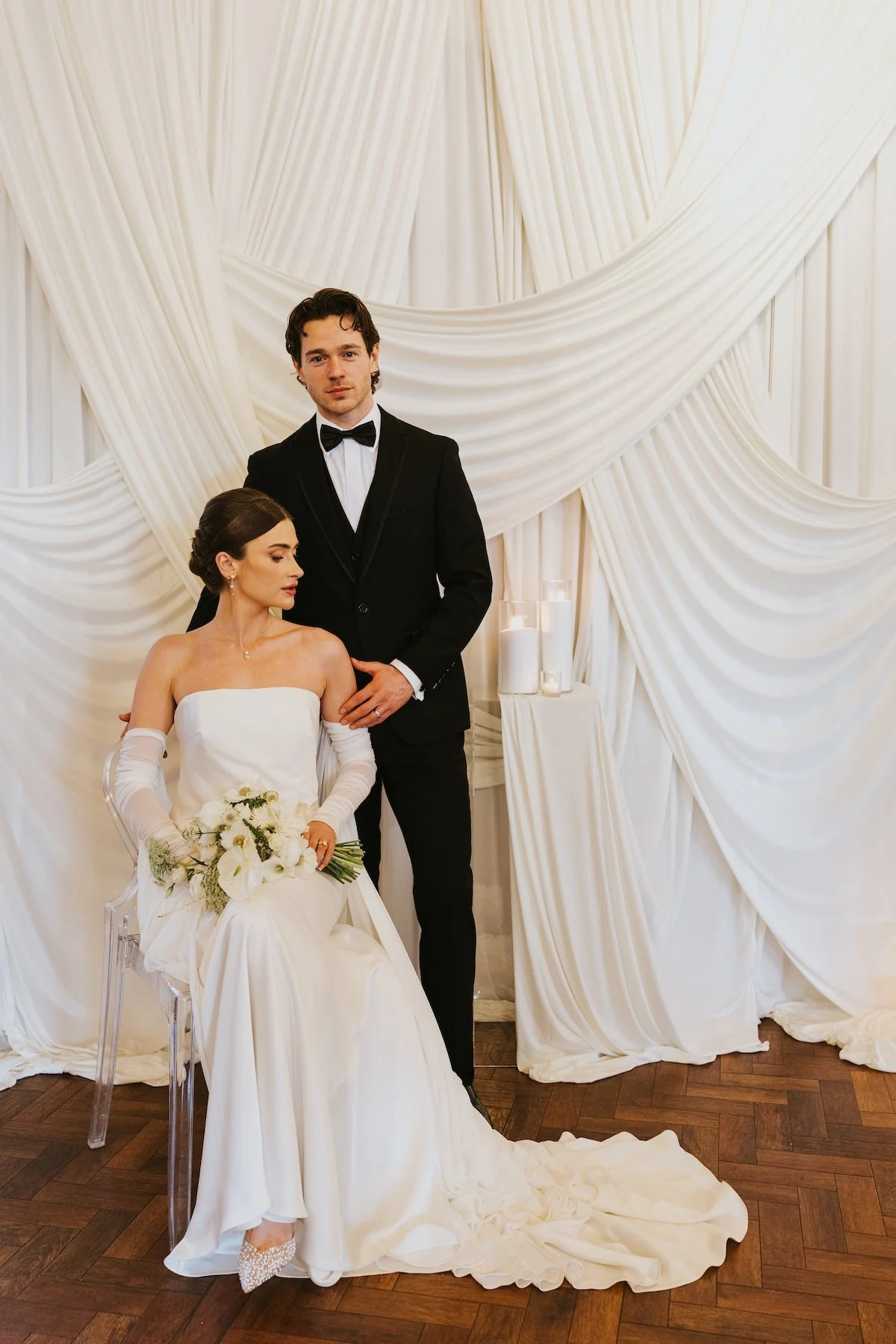 A newlywed couple dressed in wedding attire posing in front of a white draped backdrop. The bride is sitting on a transparent chair, holding a bouquet of white flowers, with her eyes closed and head slightly tilted. The groom is standing behind her, 
