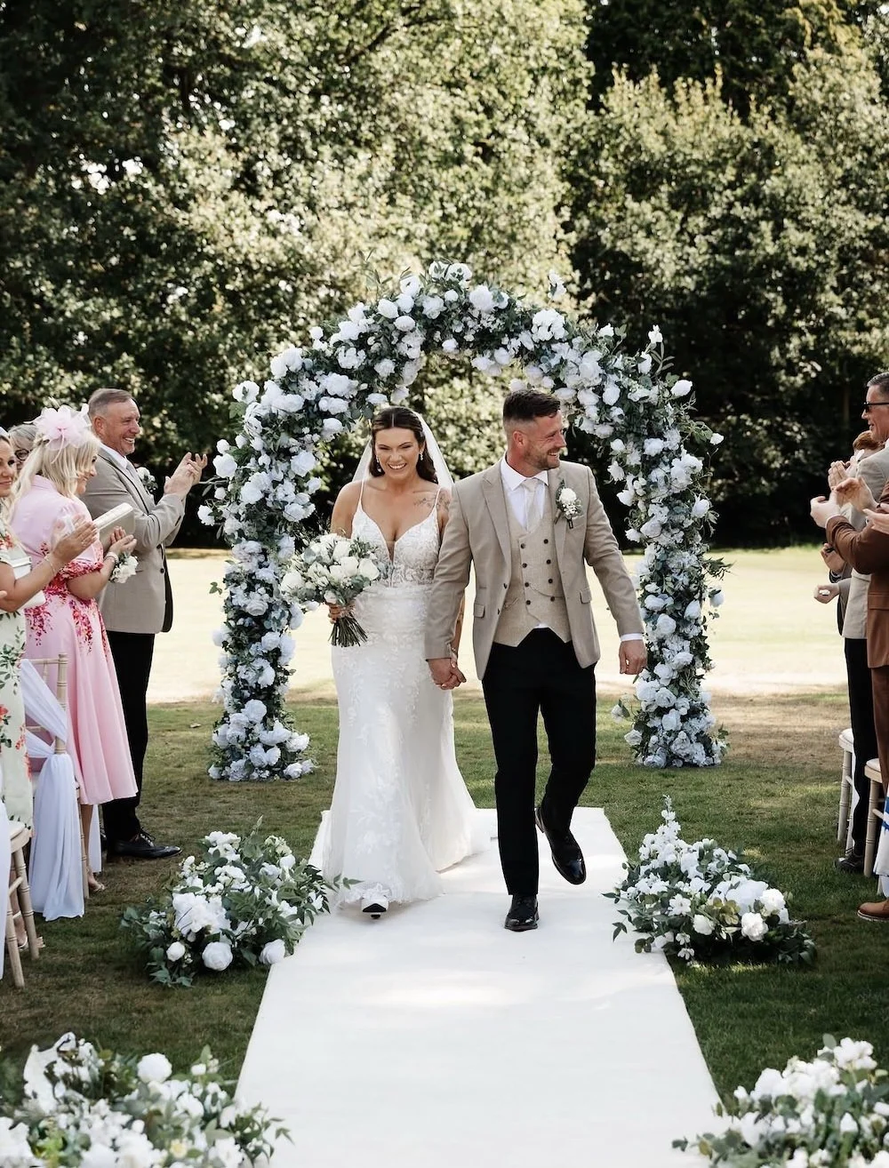 A bride and groom walk hand-in-hand under a floral arch at their outdoor wedding ceremony. The bride wears a white lace wedding dress and holds a bouquet, while the groom wears a beige suit jacket with black pants. Guests on both sides of the aisle c