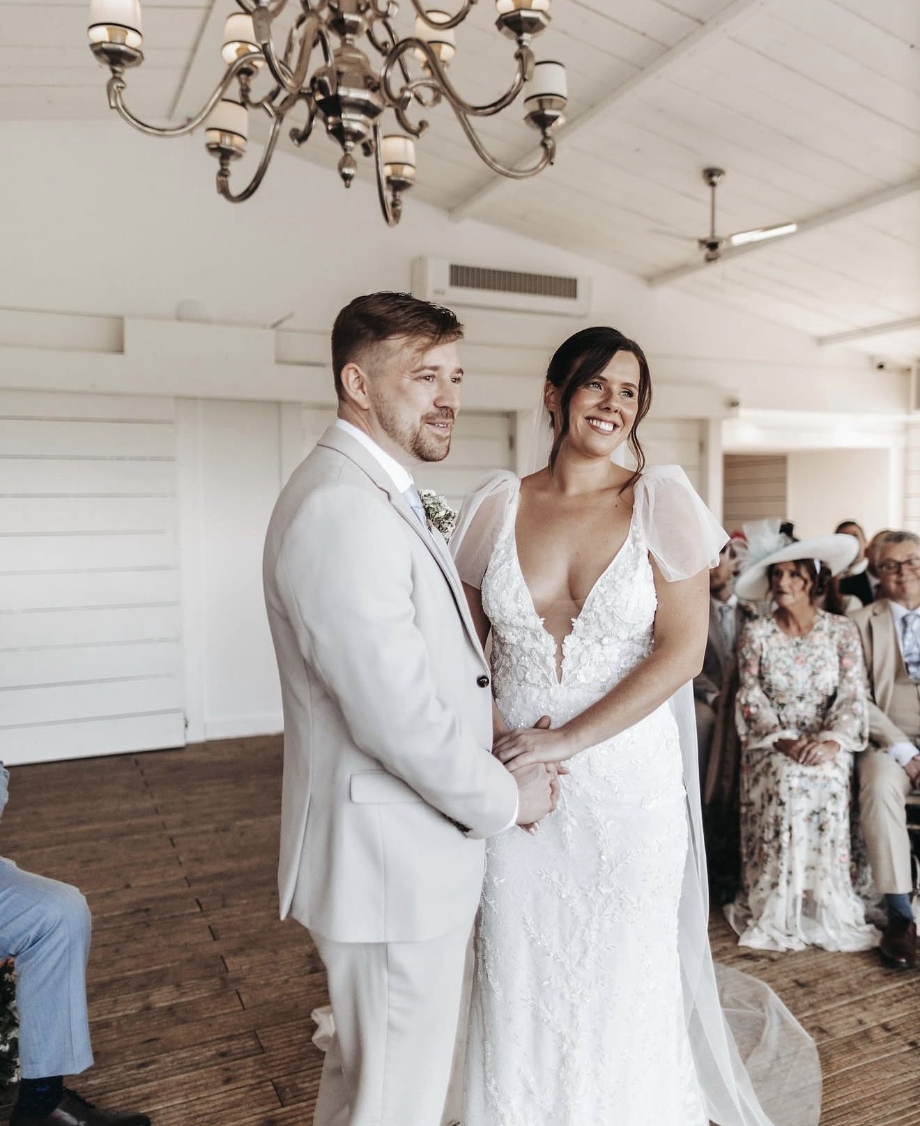 A bride and groom standing together during a wedding ceremony indoors, with guests seated behind them. The bride is wearing a white lace wedding gown with puffed sleeves, and the groom is in a light-colored suit with a white shirt and tie. The settin