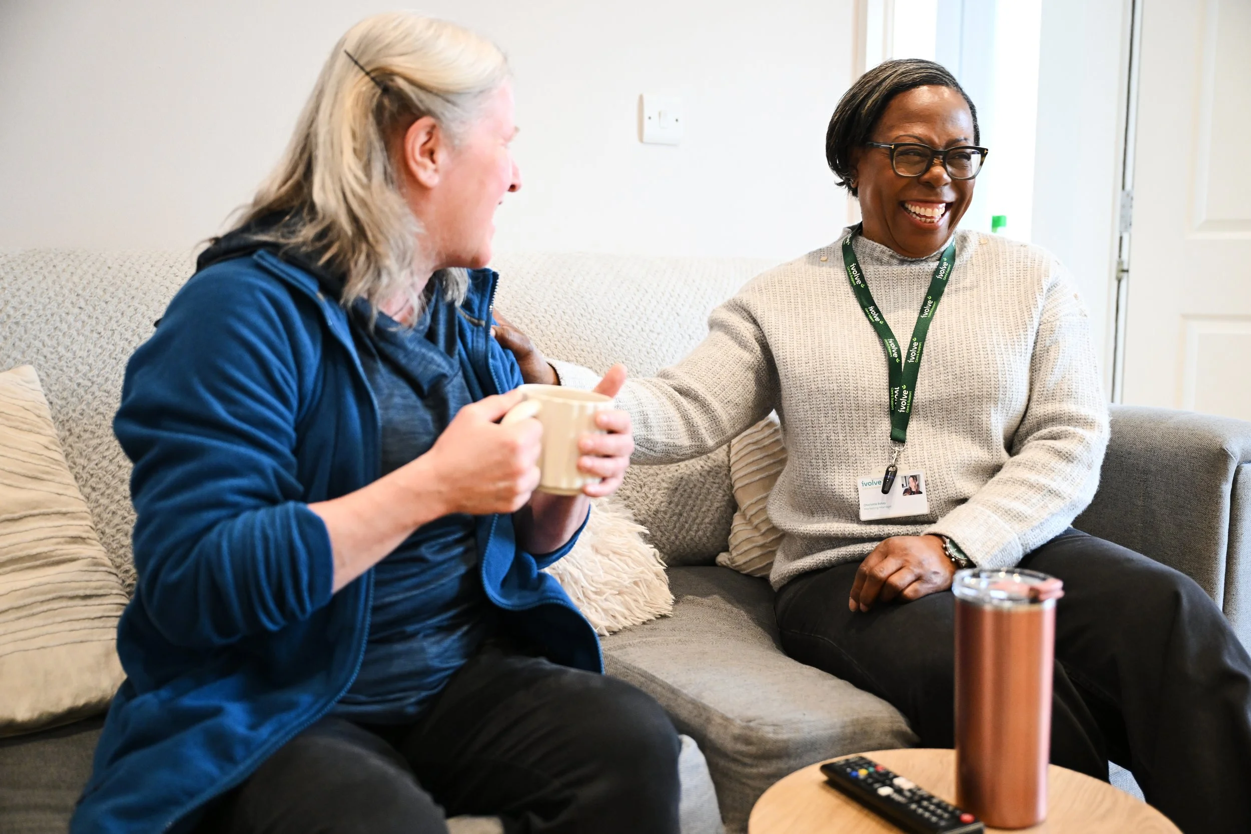 Two women sitting on a beige sofa, smiling and engaging in conversation. One woman, with platinum blonde hair and wearing a blue jacket, is holding a mug. The other woman, with short dark hair, glasses, and wearing a cream-colored sweater with a lanyard, is smiling broadly. There is a round wooden table in front of them with a remote control and a tumbler on it.