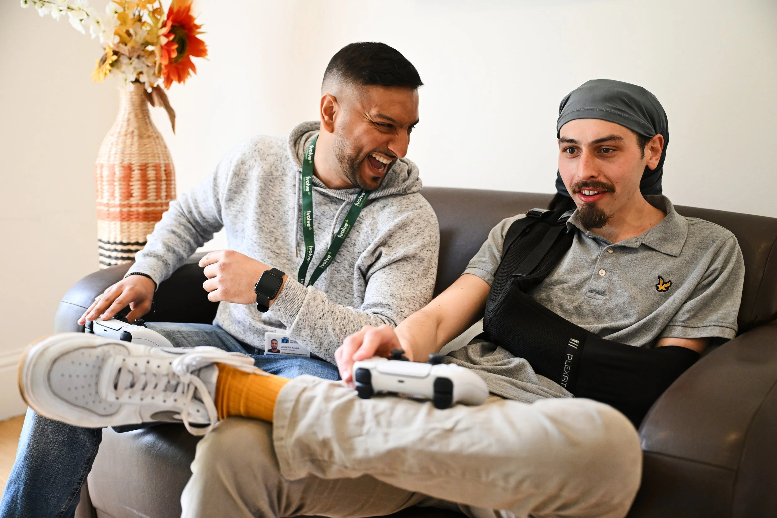 Two men sitting on a couch playing video games together, with one man laughing and the other focused, in a living room with a vase of flowers in the background.
