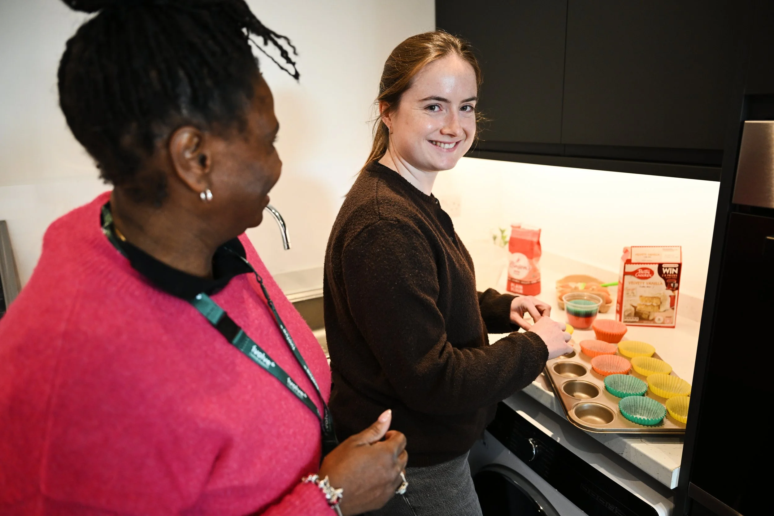 Two women standing by a countertop with colorful muffin liners and cupcake batter, one woman is smiling at the camera while the other looks at the cupcake ingredients.