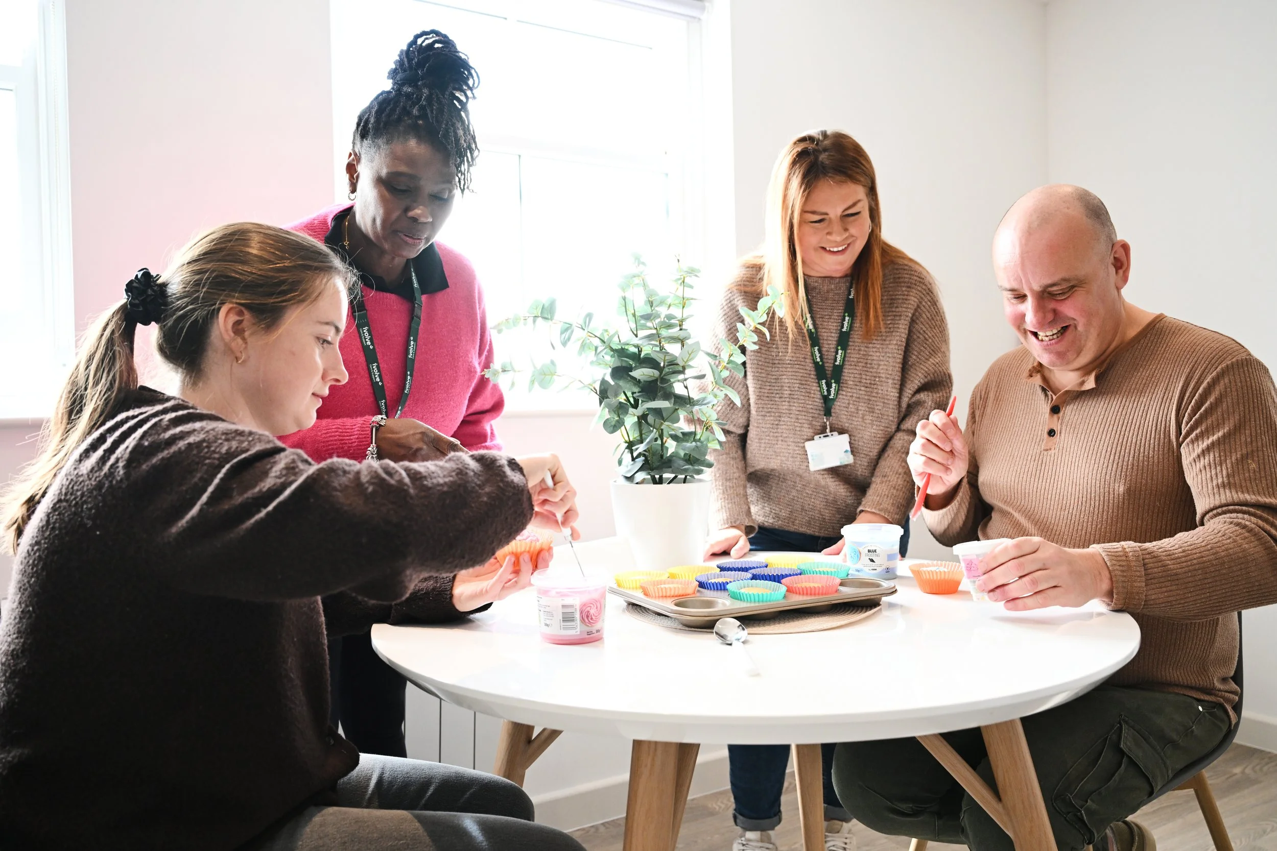 Four people gather around a round table, baking cupcakes with colorful silicone wrappers. One woman is mixing, another woman and a man are decorating cupcakes, and a young girl is holding a cupcake.