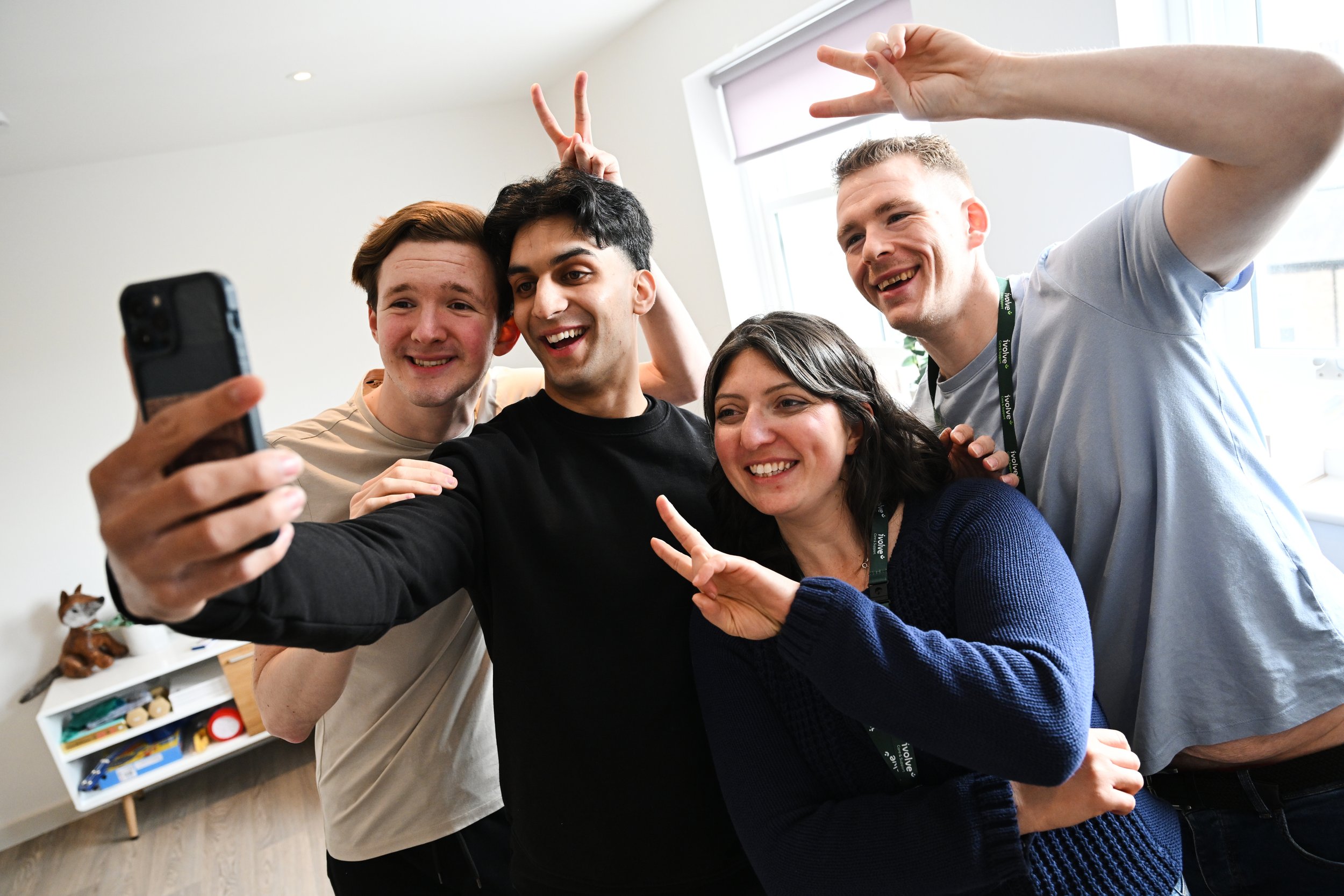 Group of five young adults taking a group selfie in an indoor setting, smiling and making peace signs.