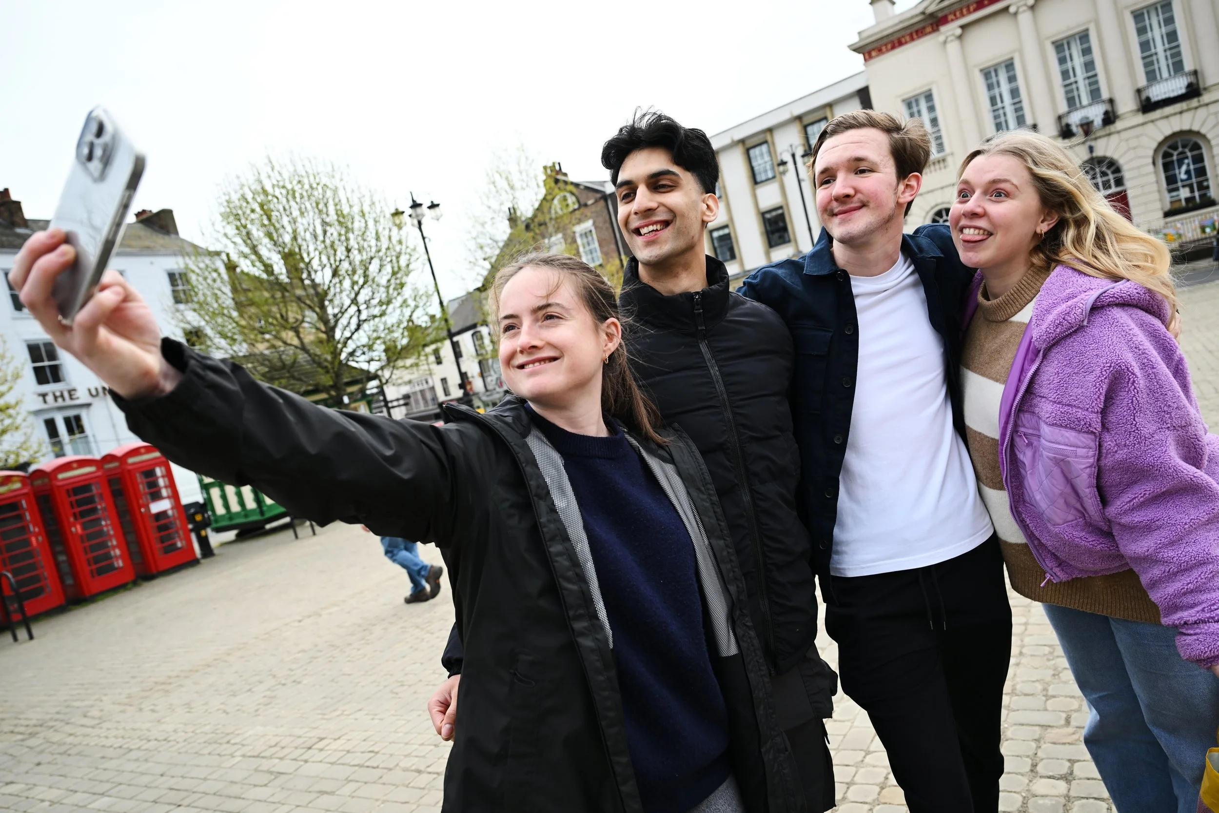 Four young people, two men and two women, are taking a selfie outdoors in a town square with historic buildings and red telephone booths in the background. They are smiling and casually dressed.