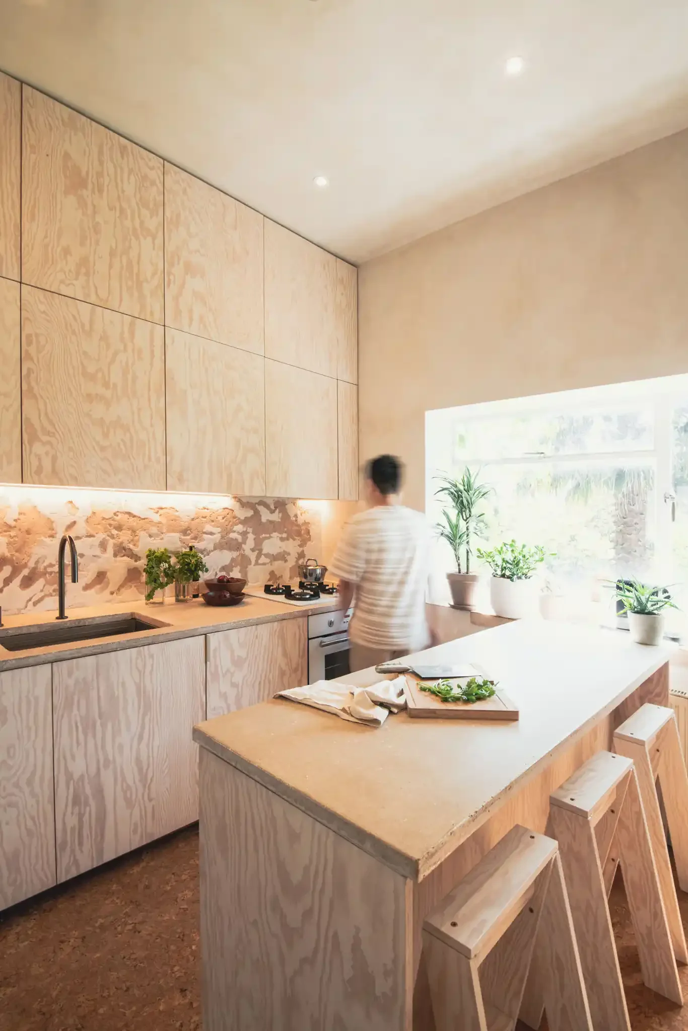 Modern kitchen with light wood cabinets, a large island with a beige countertop, and several potted plants near a window. A person is preparing food on the counter.