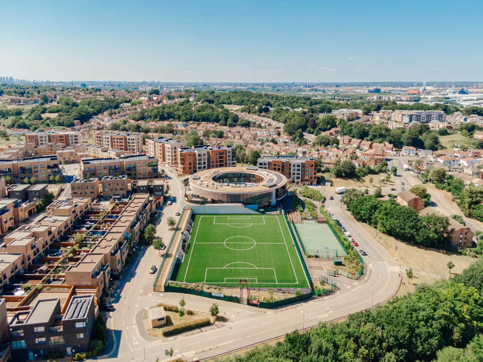 An aerial view of circular donut shaped architecture, Lime Wood Primary School in Erith, Kent. A cityscape with a soccer field at the center, surrounded by residential buildings, green trees, and roads, with a distant skyline under a clear blue sky.