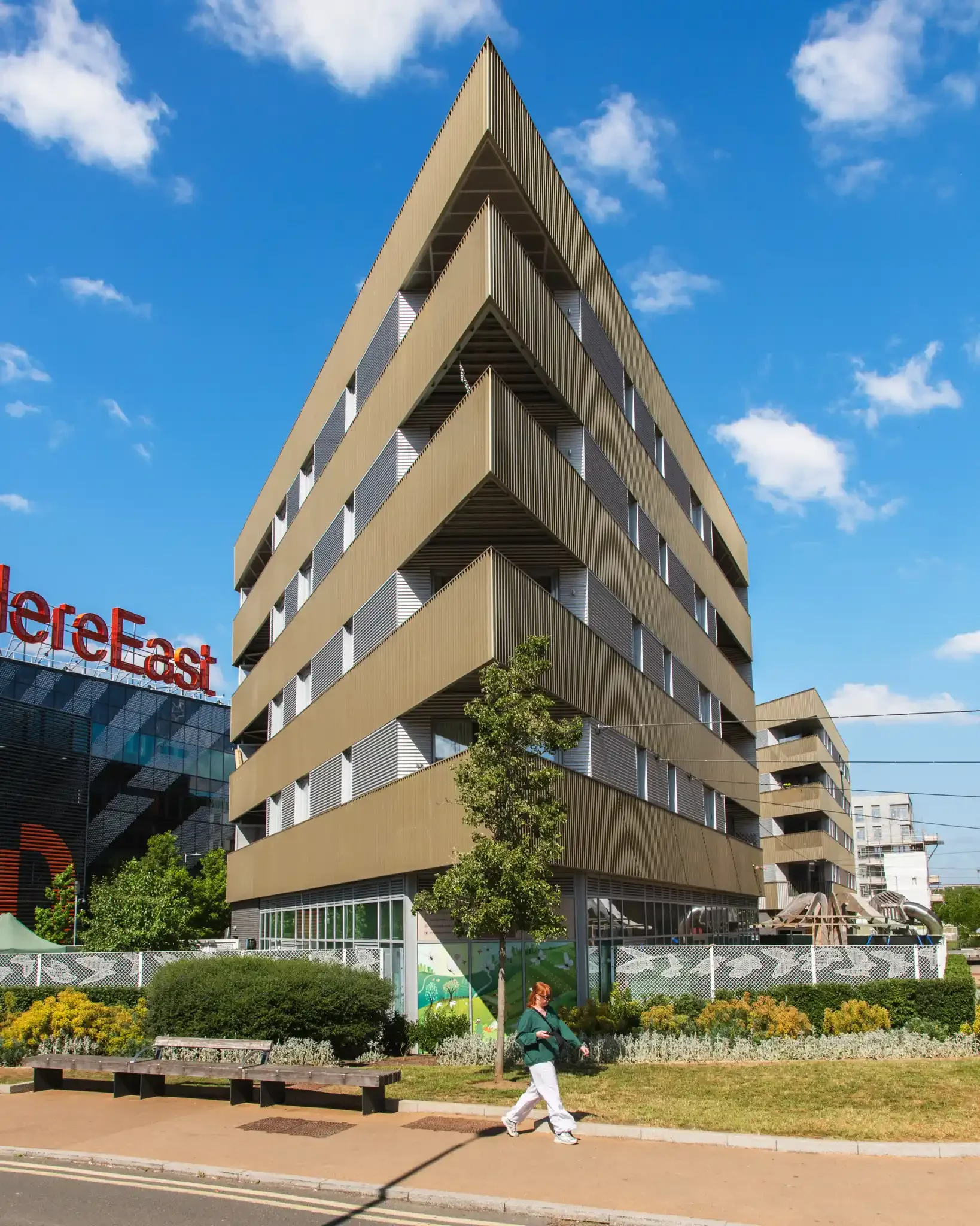 A modern multi-story building with horizontal beige slats on its facade, set against a blue sky with a few clouds, with trees and greenery at its base, and a woman walking along the sidewalk.