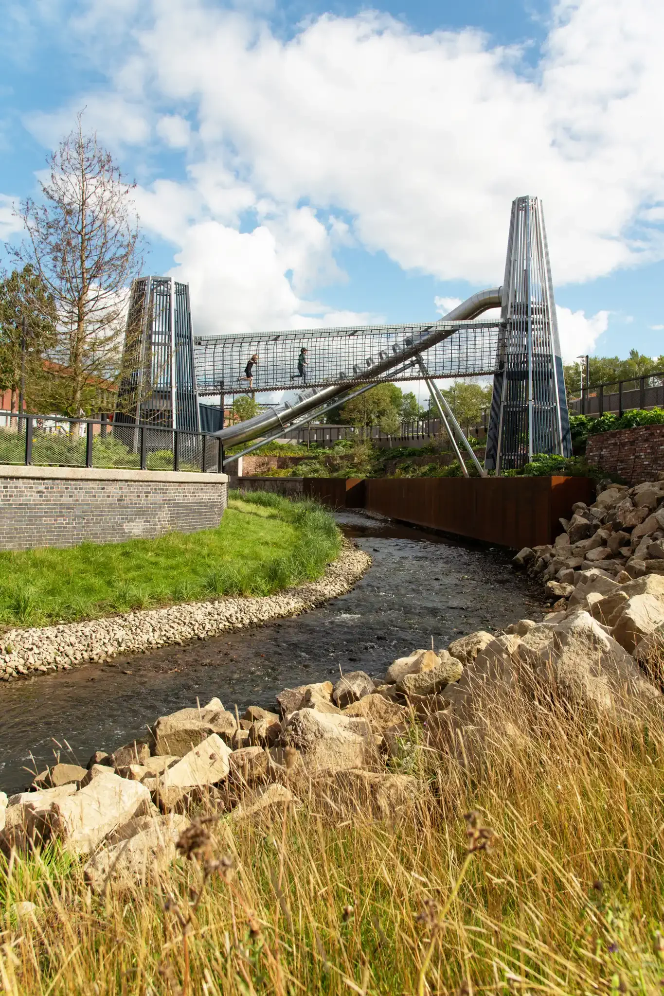 Mayfield Park playground slide towers over the River Cam. People walking on a modern, elevated bridge over a small water stream, with green grass, rocks, trees, and a partly cloudy sky in the background.