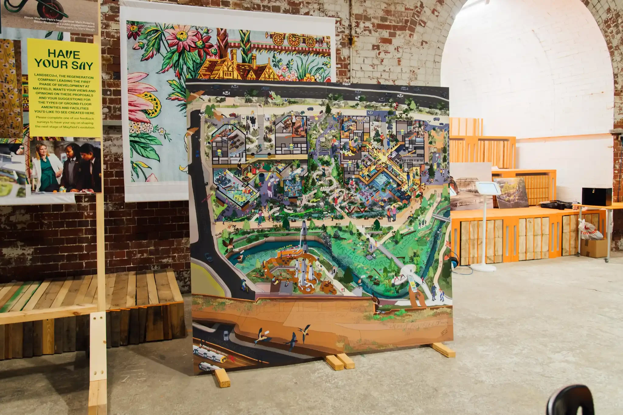 A detailed, colorful architectural site plan or model of a community or park, displayed on an easel, with a brick wall and some artwork in the background.