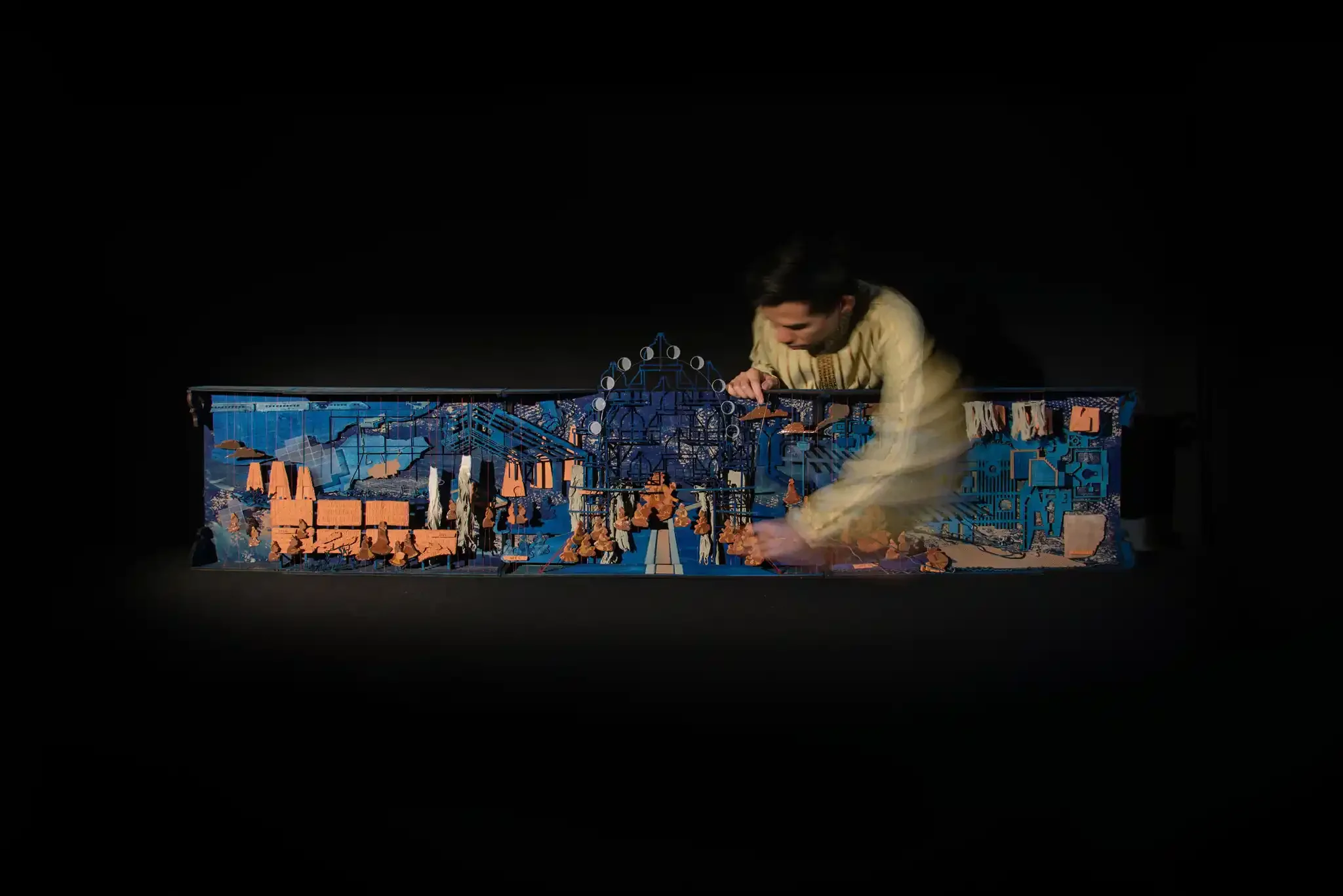 A woman wearing traditional clothing kneels beside a miniature model landscape with buildings, trees, and a Ferris wheel, set against a black background.