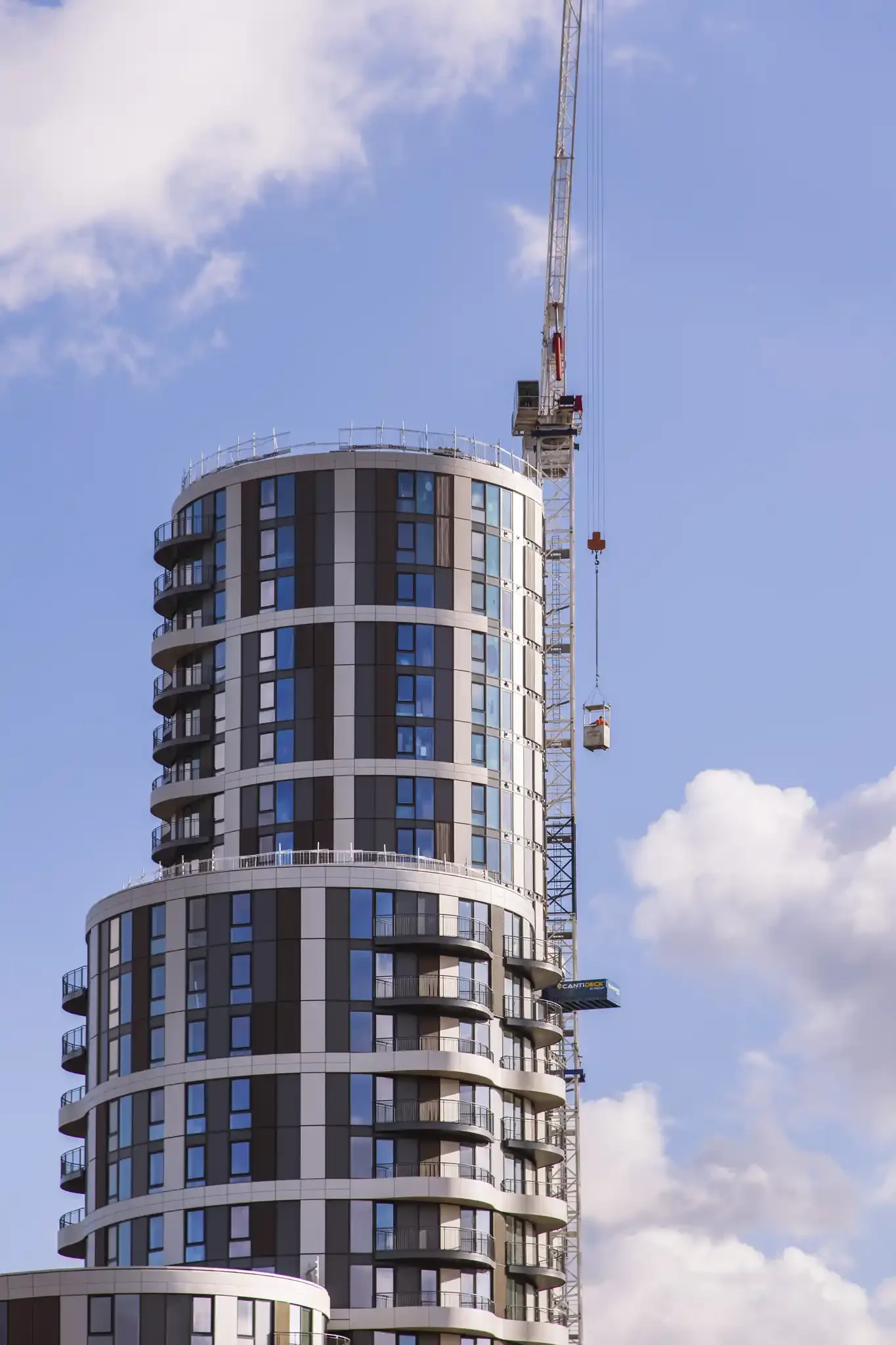 A high-rise building under construction with a tower crane on top, set against a blue sky with scattered clouds.