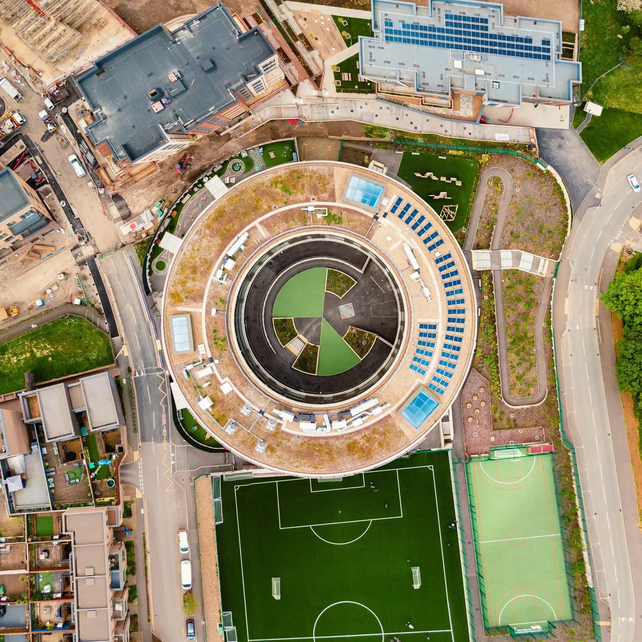 An aerial view of a cityscape featuring a school with a soccer field, surrounding buildings, parking areas, and roads, with construction and greenery nearby.
