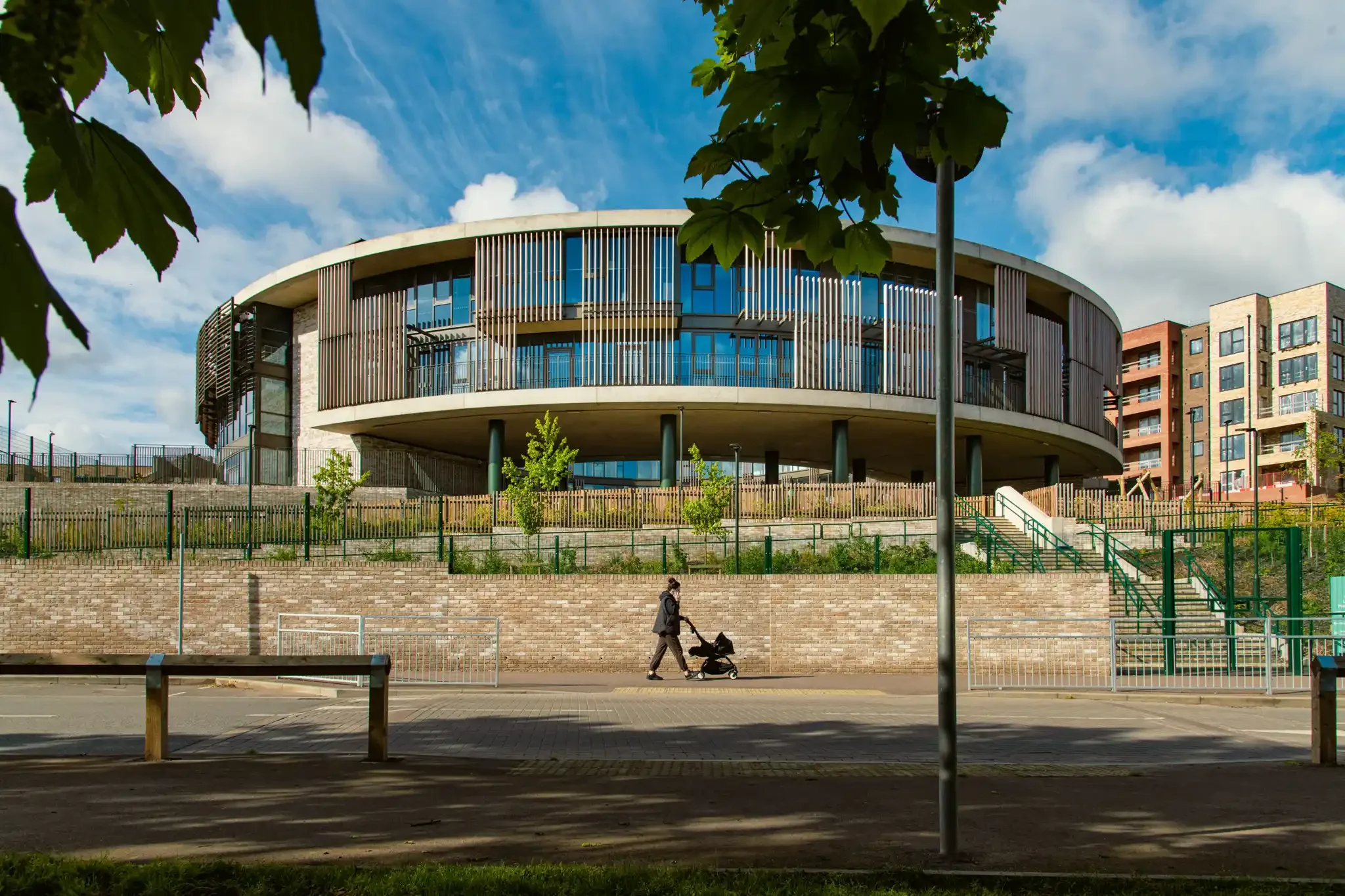 A curving modern building with large glass windows and vertical slats, situated above a brick retaining wall with stairs and a fenced area, under a partly cloudy blue sky. In the foreground, a person pushes a stroller on the sidewalk.