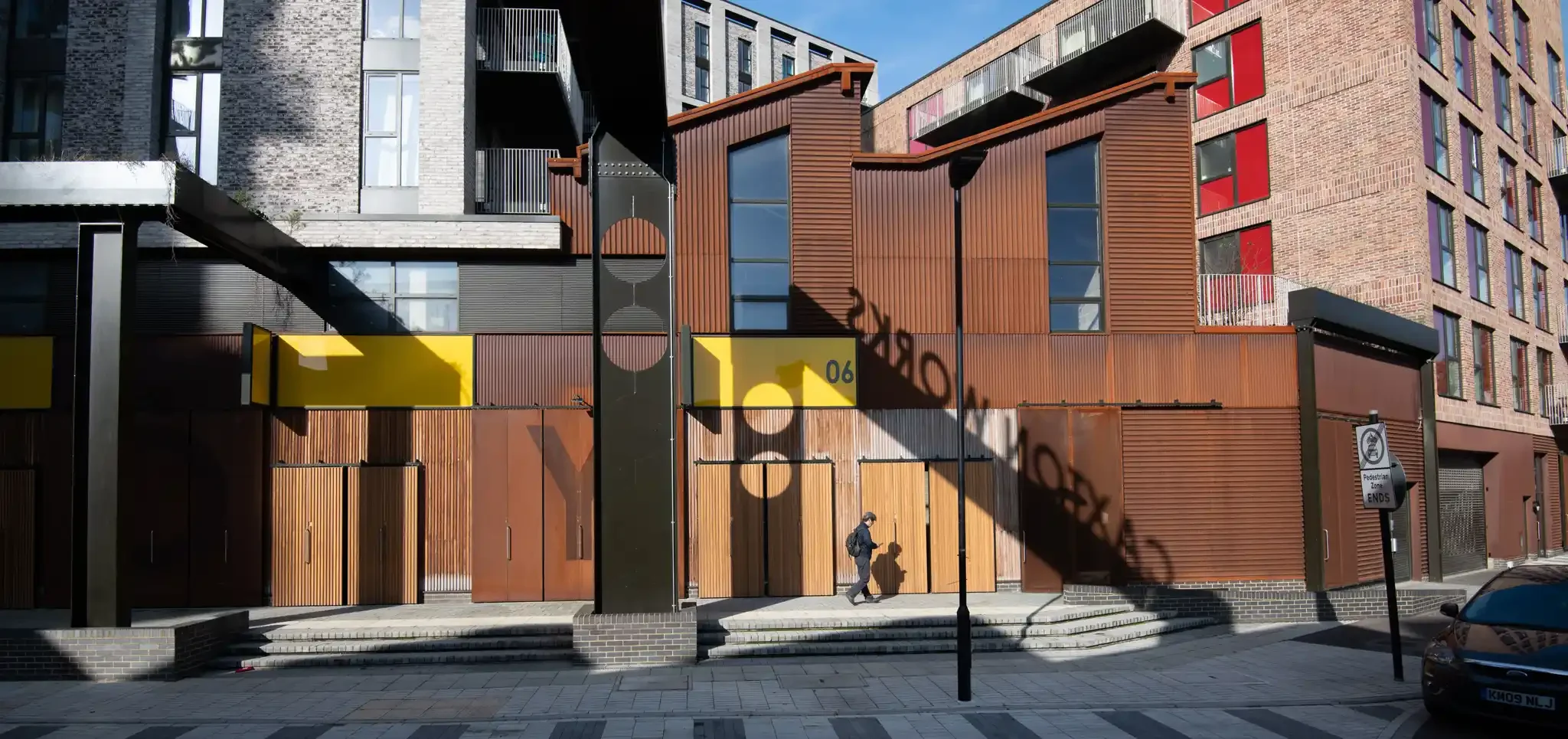 Modern multistory building with brick, metal, and wood exterior, large windows, and balconies, with pedestrians walking past and a street sign indicating pedestrian zone.