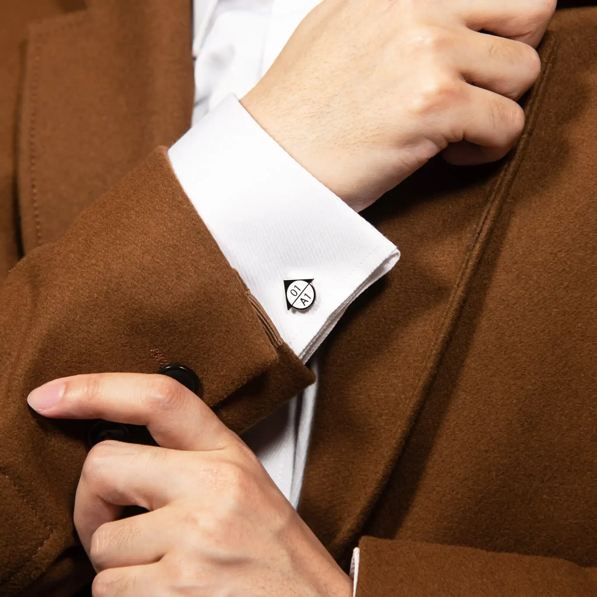 Close-up of a person's hand resting on their chest, wearing a brown coat, a white shirt, and a black button, with a visible white clothing tag.