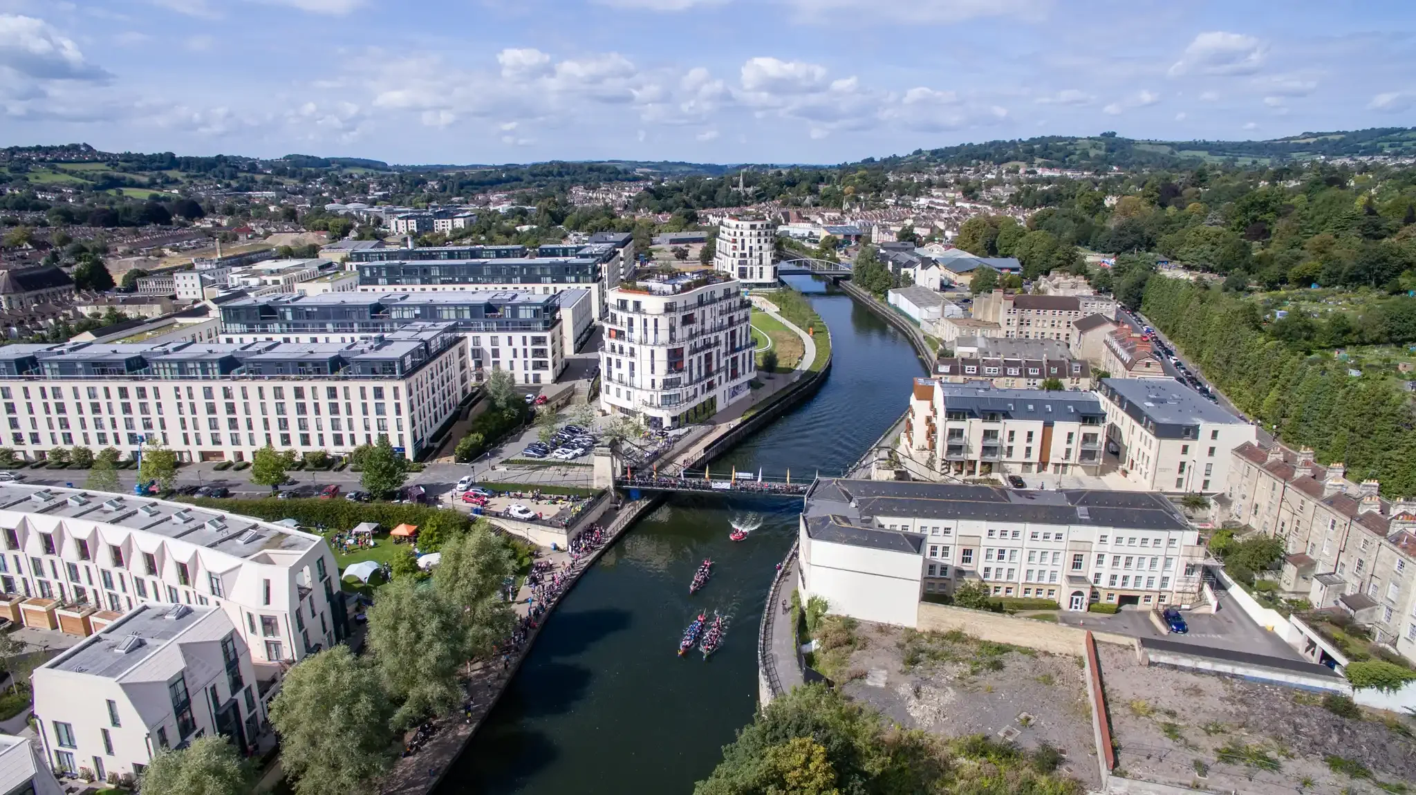 Aerial view of a cityscape with modern buildings along a river, with boats and a crowd gathered on a bridge, surrounded by greenery and hills in the background.