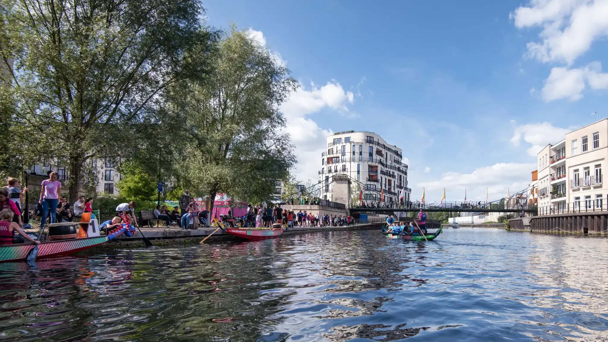 People dragon boating on a canal with modern buildings and a bridge in the background, trees on the left, and a blue sky with clouds.