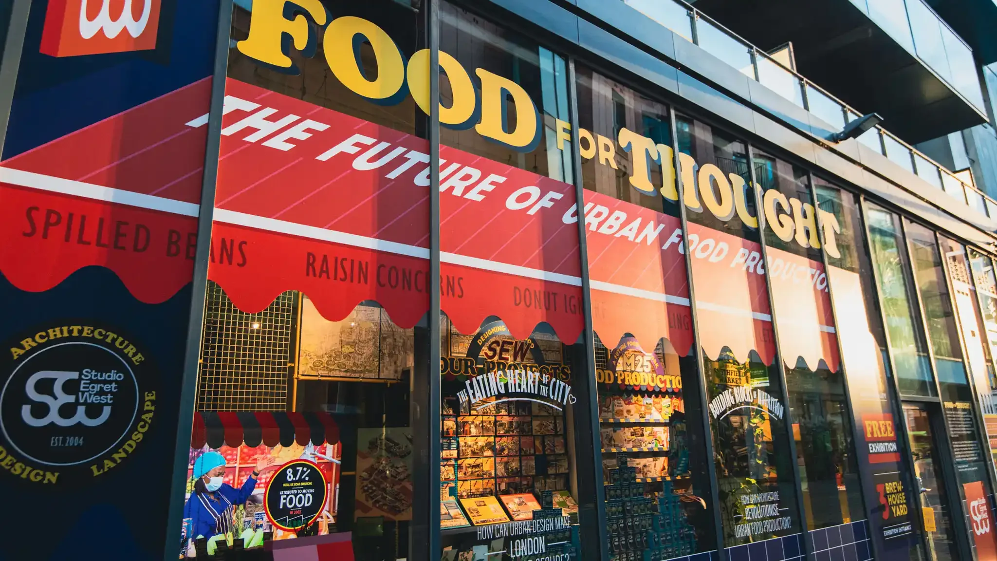 Exhibition vinyl storefront creative graphic design. Storefront with large yellow letters spelling 'FOOD FOR THOUGHT' and a red awning below with names of food items like 'spilled beans' and 'raisin cones.'