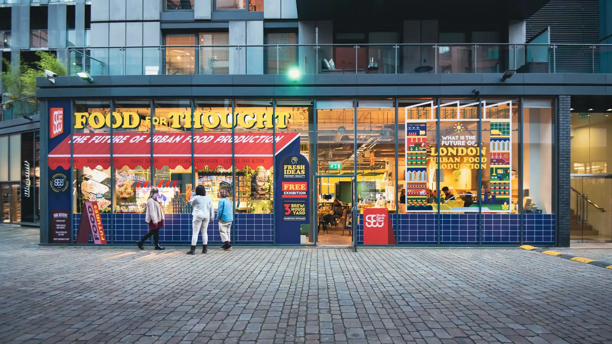 A modern glass-fronted building with a food market inside, labeled 'Food for Thought' and promoting urban food production, with a few people walking in front of it.