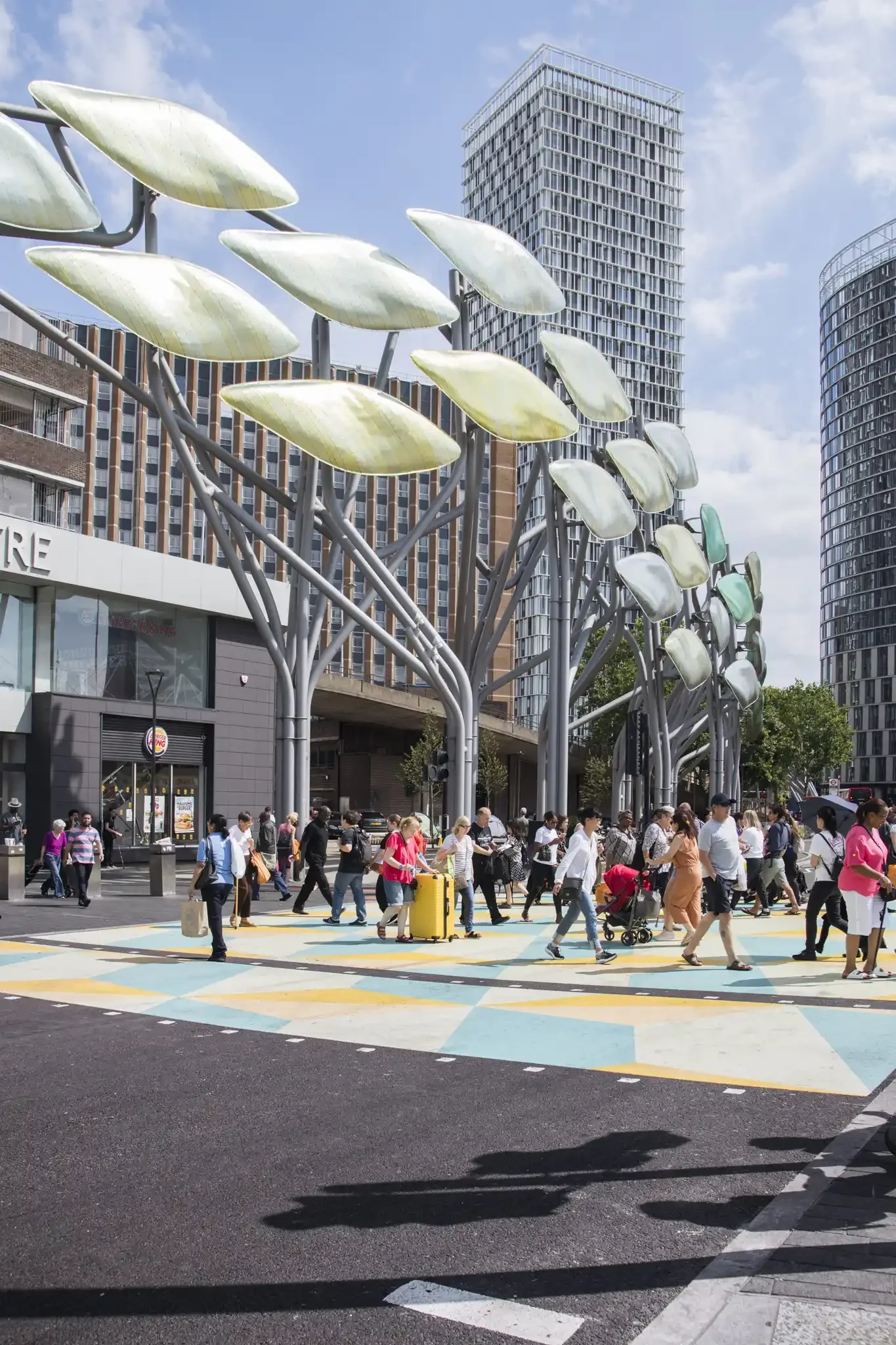 A busy city street scene with many pedestrians crossing the street. In the background, there are modern high-rise buildings and unique metal sculptures resembling tree branches with large leaves.