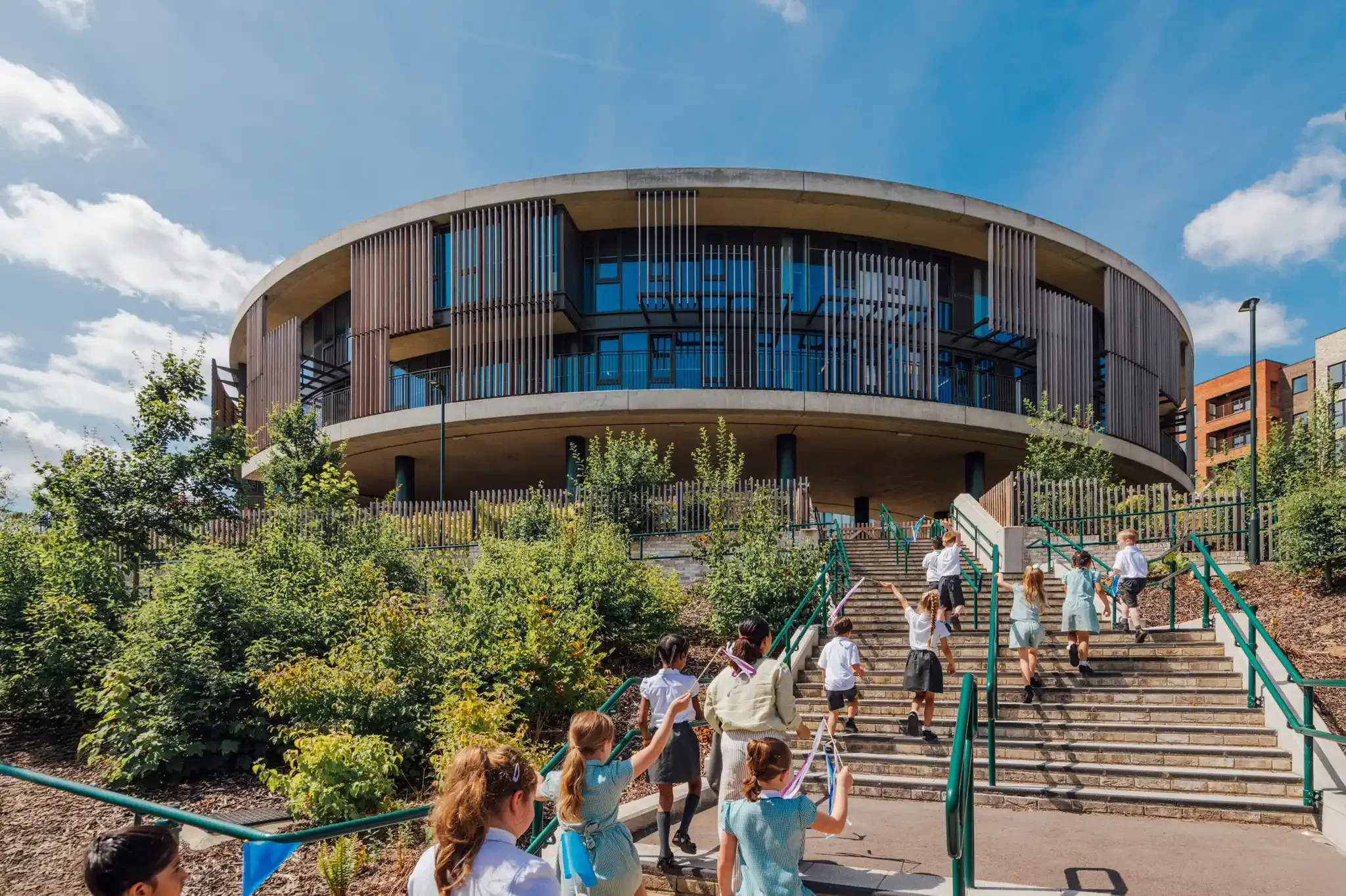 A group of children in school uniforms walking up outdoor stairs towards a modern, circular building with vertical wooden slats and glass windows, with trees and a blue sky overhead.