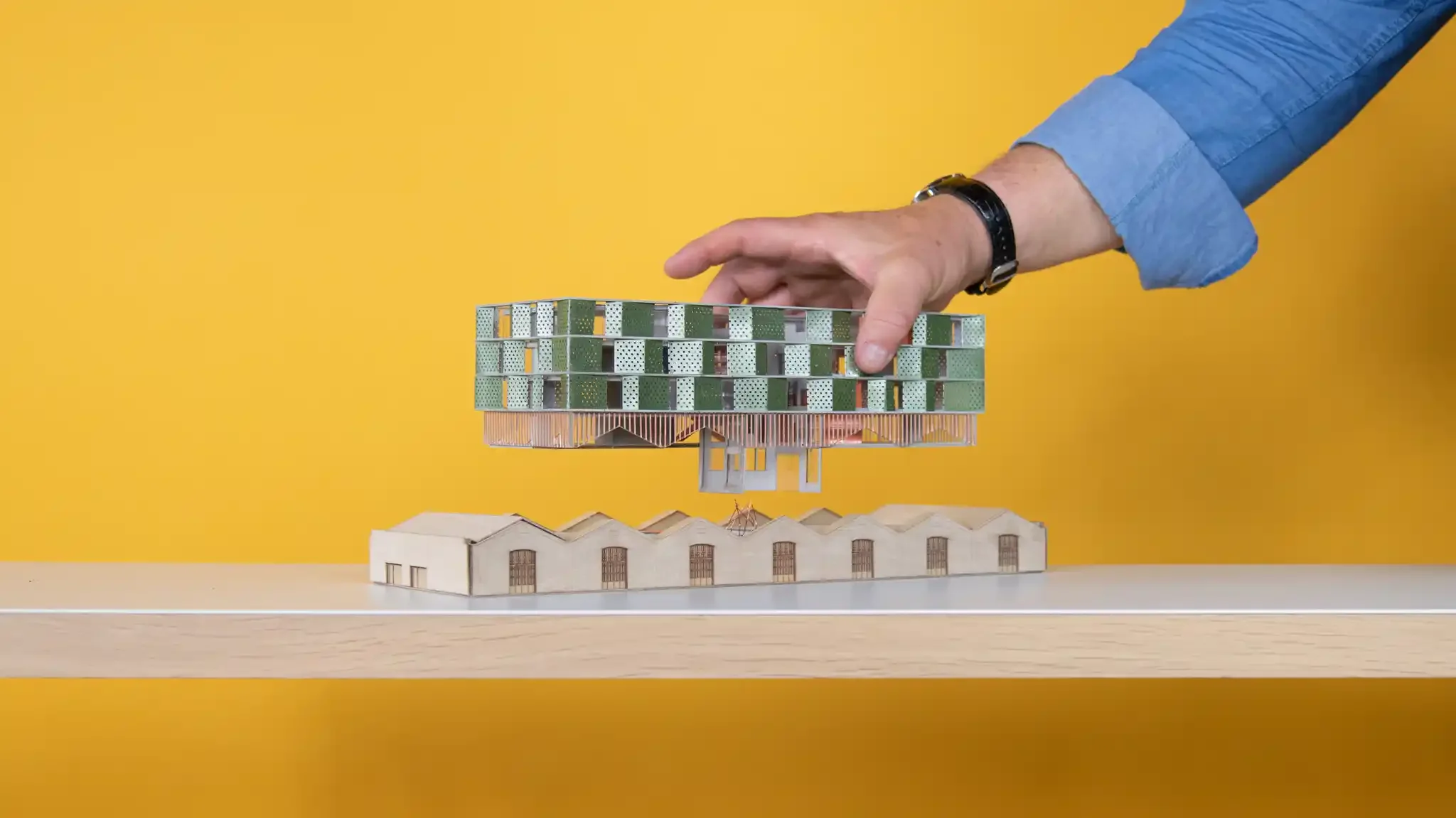 A person's hand adjusting a detailed architectural model of a modern building with a yellow background.