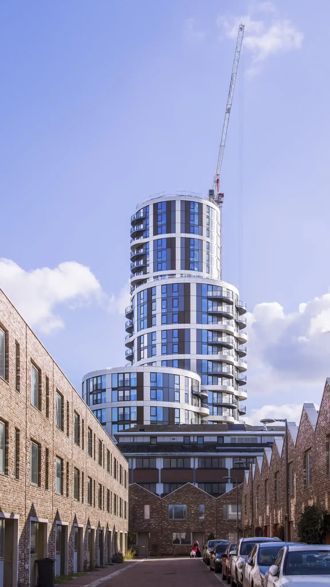 A modern high-rise building with a unique curved design under construction, surrounded by older brick buildings and parked cars.