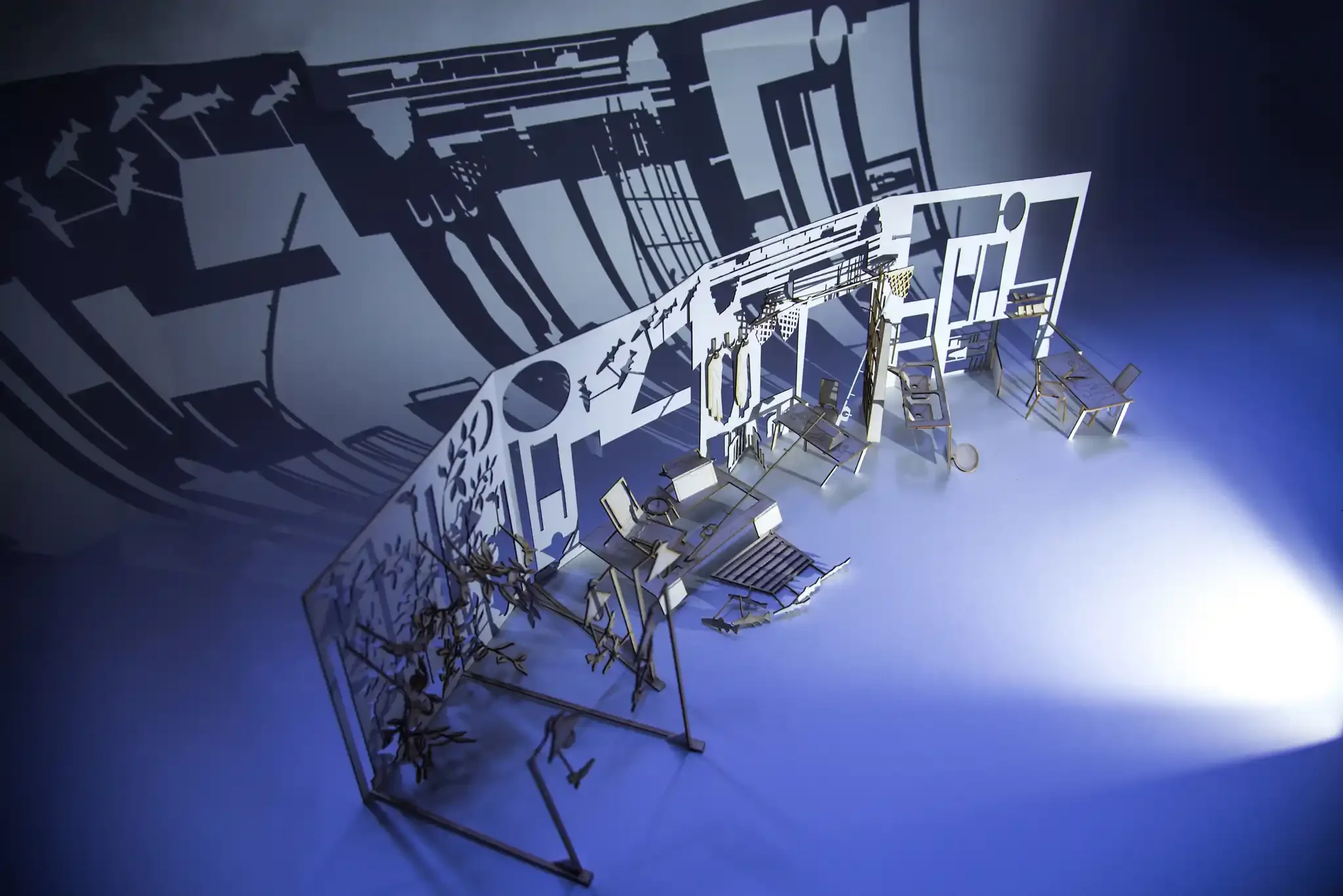 Shadow of a house silhouette with furniture and outdoor objects cast on a wall, with small tables and chairs arranged on the ground