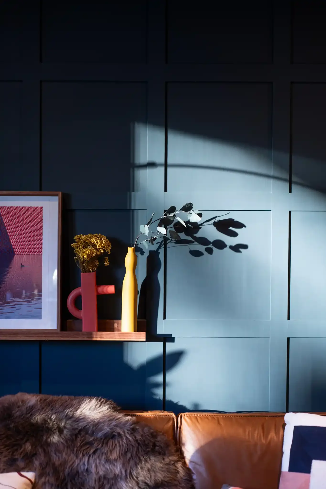 Interior of a room with a dark blue paneled wall, a shadow cast from a vase with white leaves, a framed picture with red and white pattern, a yellow vase, a pink vase, and a fur blanket on a leather couch.