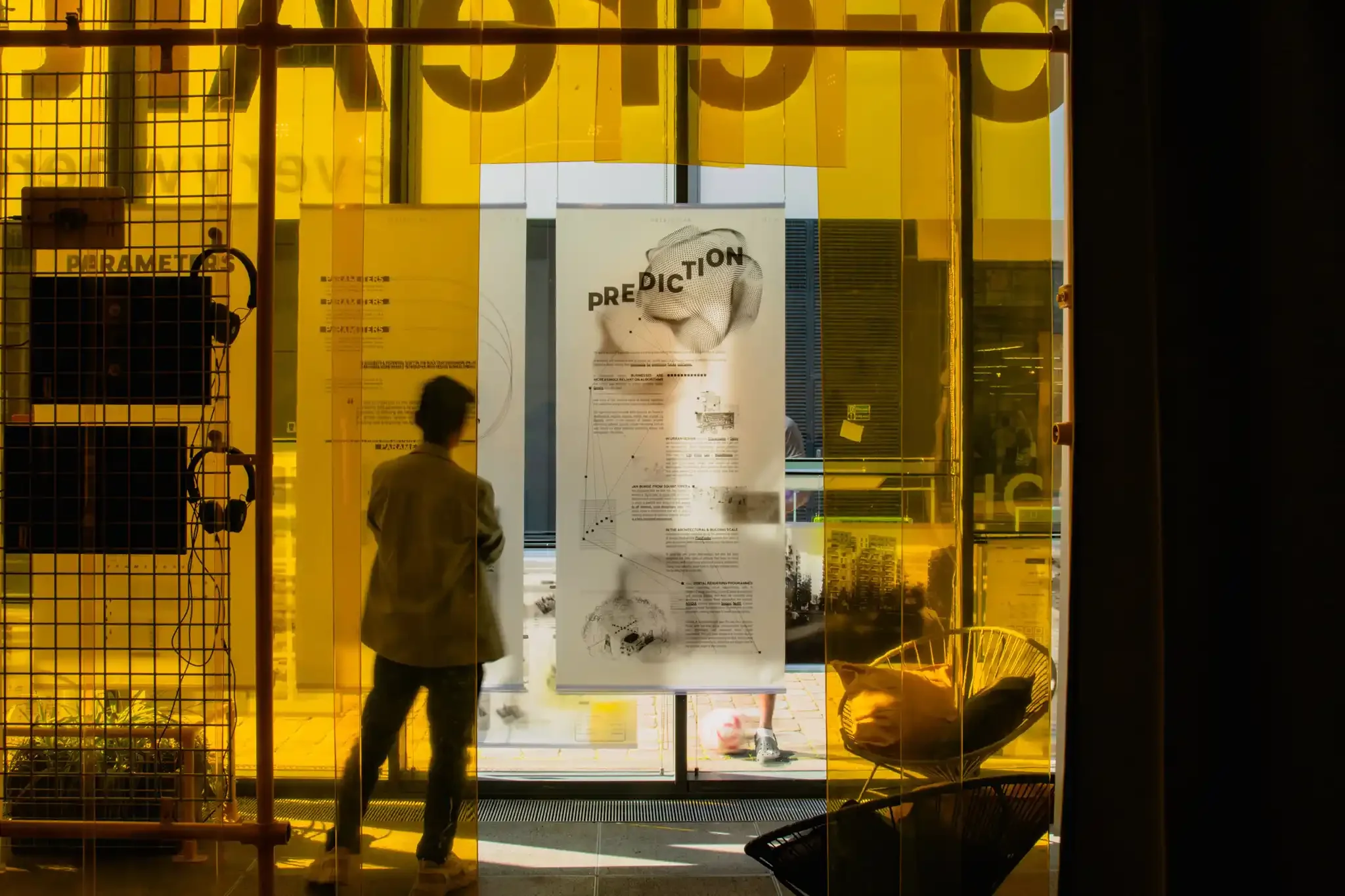 View from inside a building with a yellow tinted butchers plastic flaps, used in a exhibition design, showing a person walking by a poster with a large illustration and the word 'PREDICTION'.