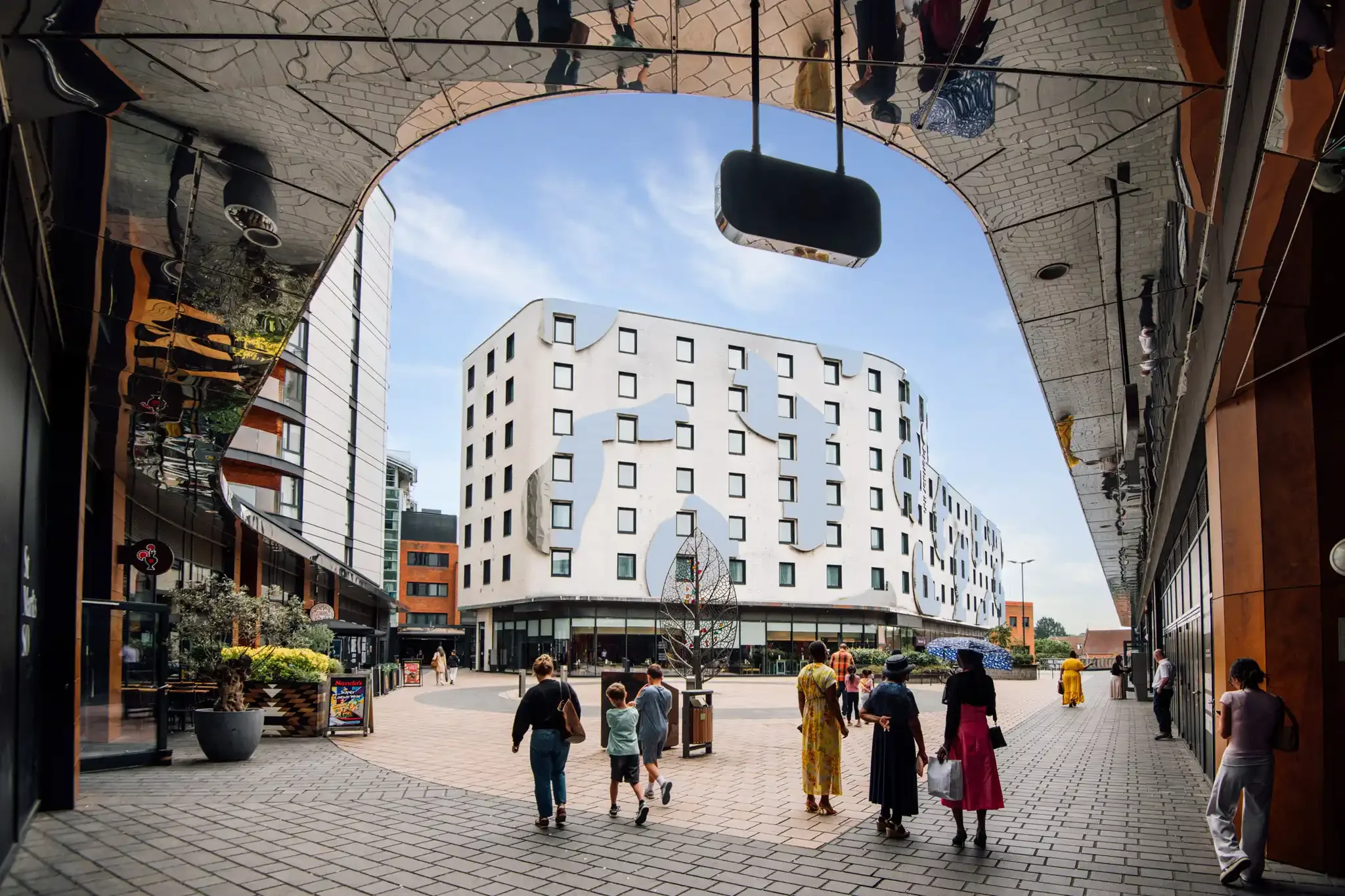 People walking in a shopping area with modern buildings and a patterned reflective soffit walkway overhead. St Marks Square in Bromley. The scene includes storefronts, outdoor plants, and a clear sky.