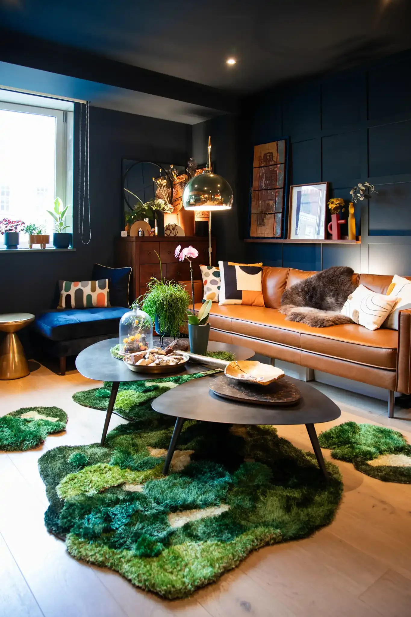 Living room with blue walls, brown leather sofa with pillows, green patterned rug, black coffee tables with decorative items, plants, window with plants on sill, framed pictures on shelf, modern lighting, and a furry blanket on sofa.