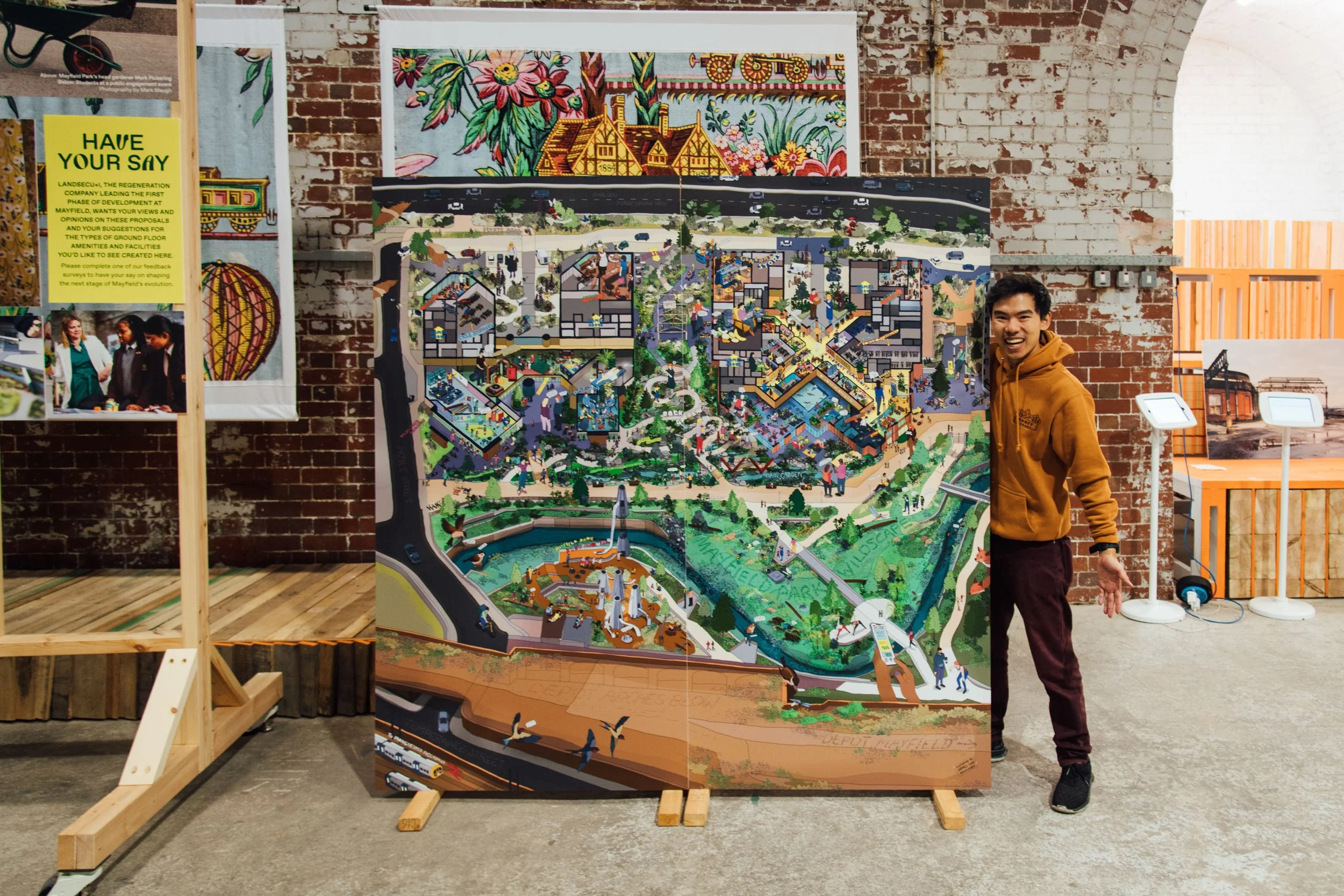 A large, detailed map of Mayfield Park in Manchester is displayed on a stand, with a smiling young man in a brown hoodie standing next to it. The large drawing is a detailed masterplan illustration of the future neighbourhood, drawn in isometric.