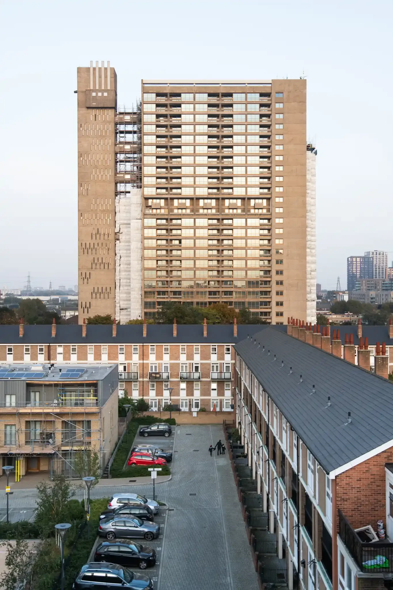 View of a high-rise building under construction with scaffolding, surrounded by lower residential buildings and a parking lot with parked cars and two people walking.