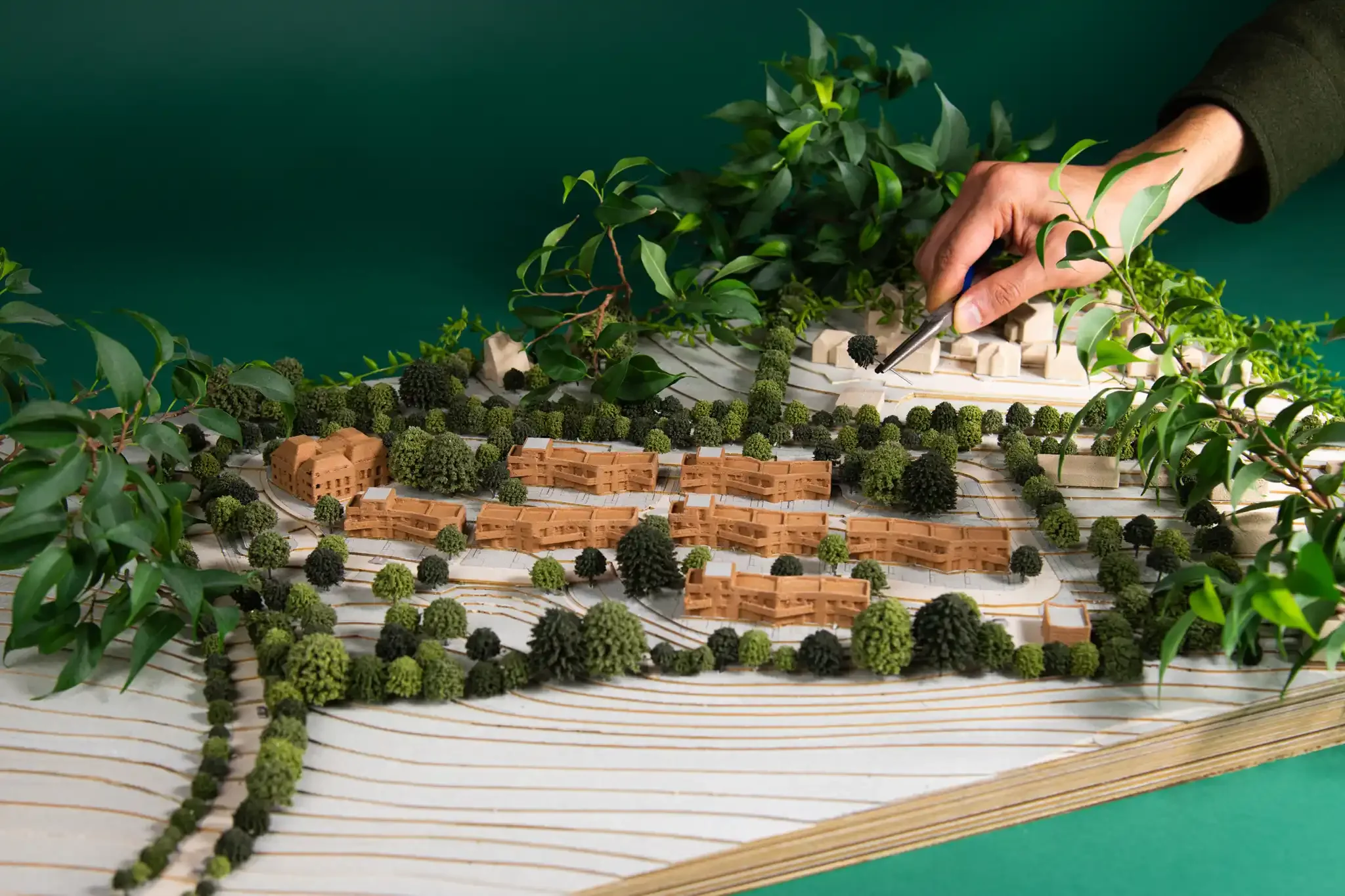 A person working with a small trowel on an architectural model of a landscape, including trees, buildings, and pathways, with greenery surrounding the scene. Stylised staged product photography of a architectural model.
